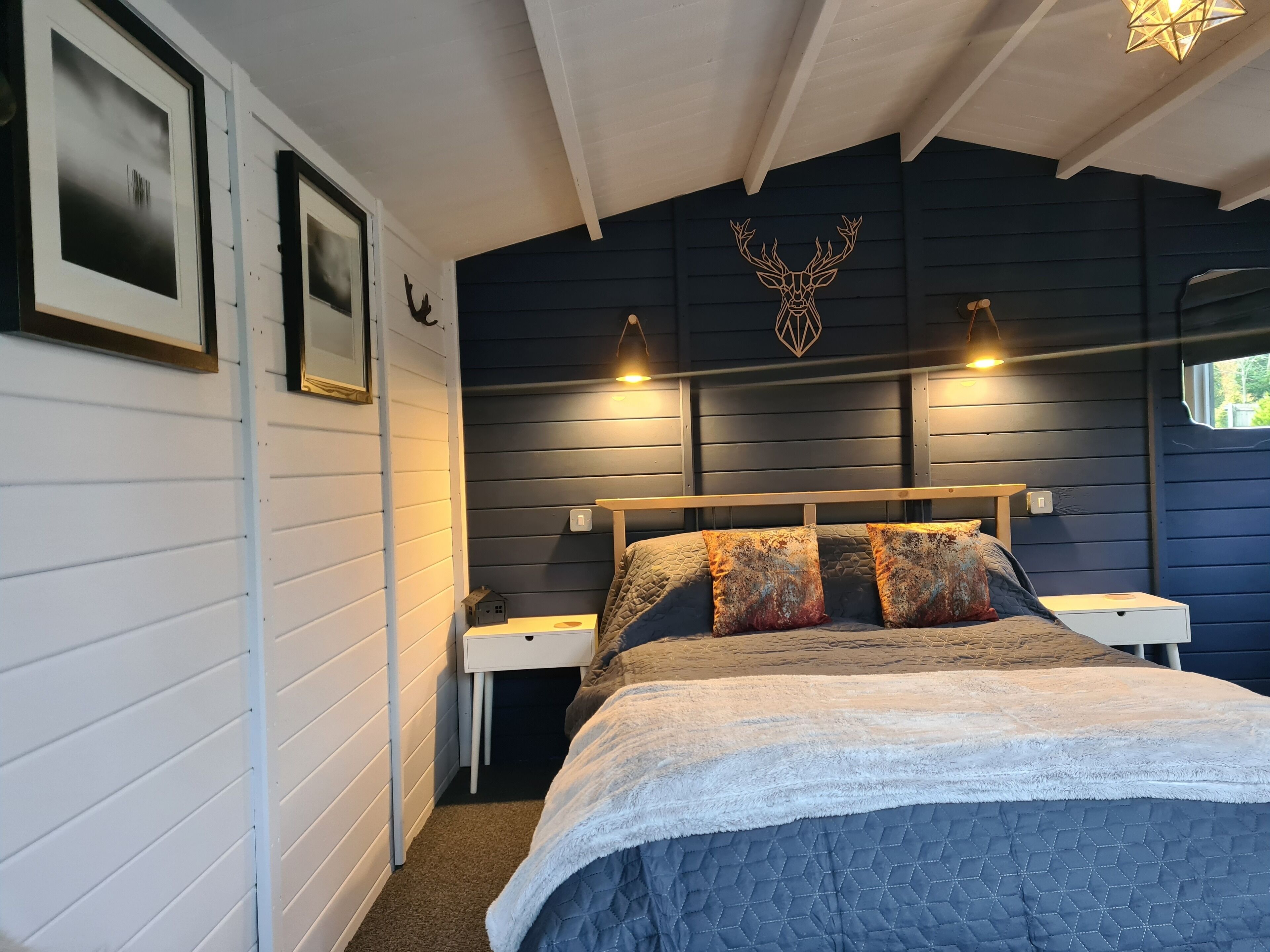 A bedroom with a bed, two bedside tables, a nightstand, a wall-mounted light fixture, and a window.