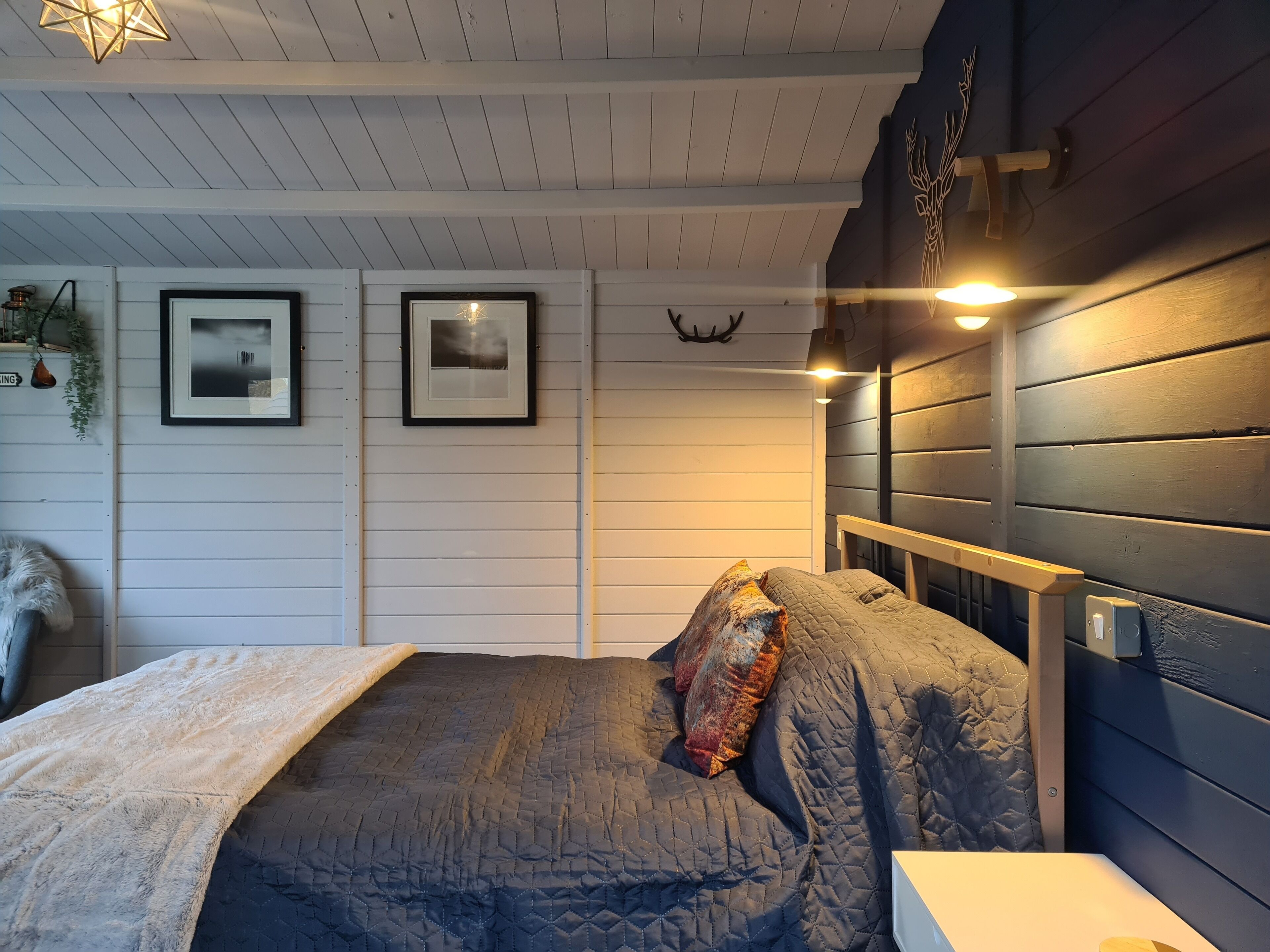 Une chambre à coucher avec un lit, une table de chevet, une lampe fixée au mur et des tableaux encadrés.