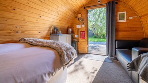 A cozy cabin room with a bed, a sofa, a small kitchenette, and a door leading outside. - The Little Hide - Adult Only Camping Pods (York)