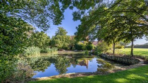 A serene pond surrounded by lush greenery and trees, with a stone wall and a wooden bridge in the background. - The Little Hide - Adult Only Camping Pods (York)