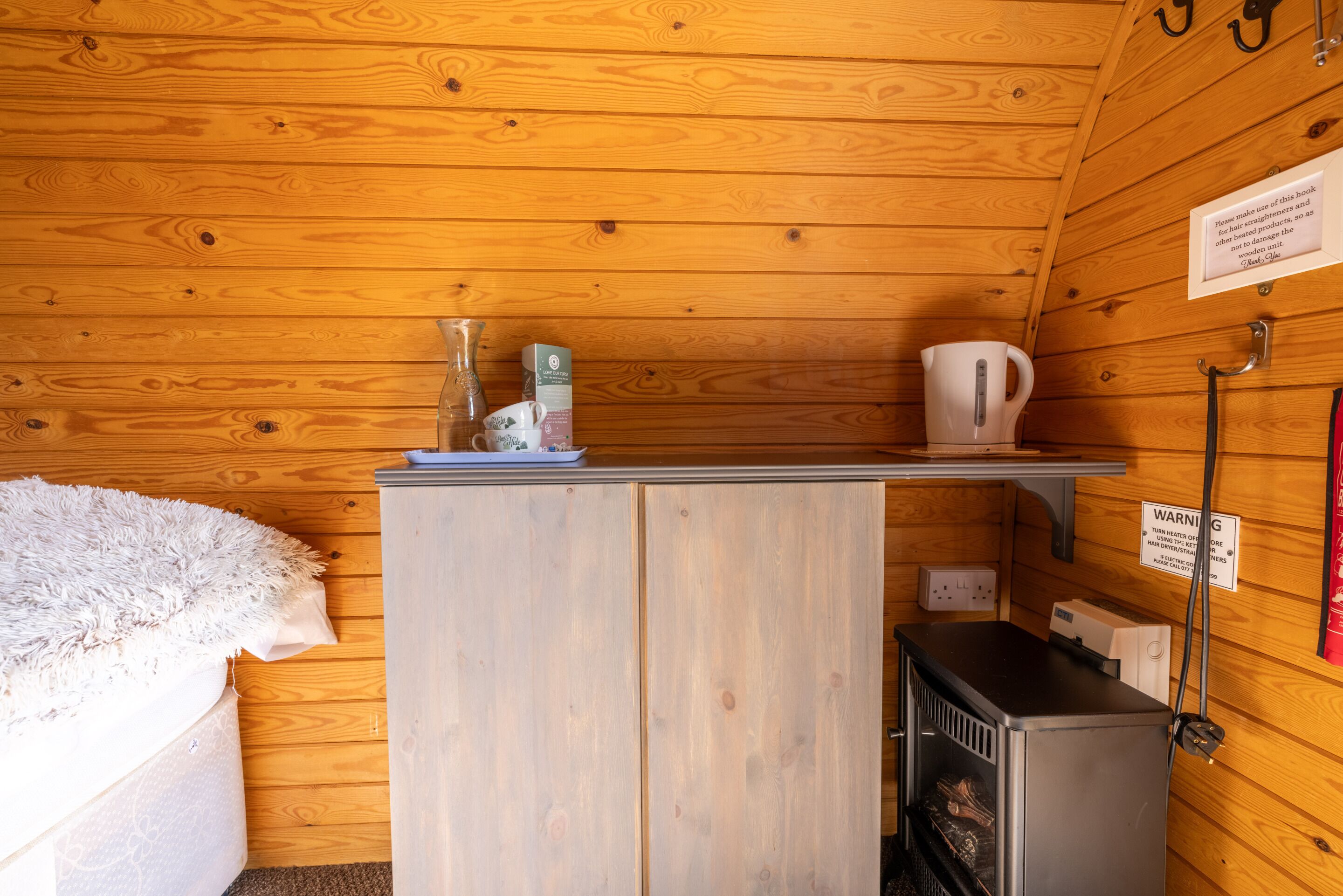 Interior de una cabaña de madera con una cama, una repisa con una tetera y una caja, un calentador y un cartel de advertencia.