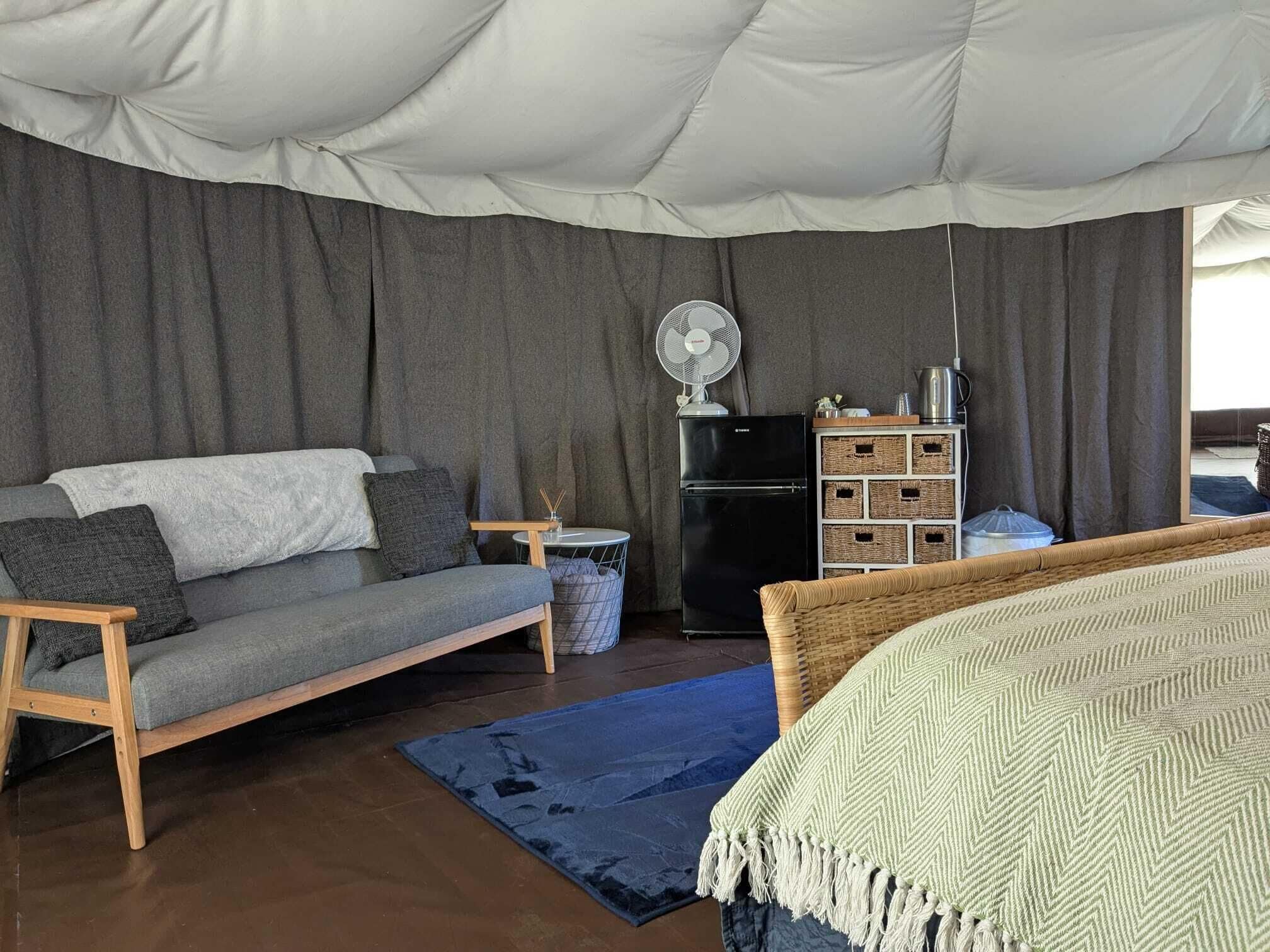 A cozy room with a grey sofa, a small round table, a black refrigerator, wicker baskets, and a bed with a patterned blanket.
