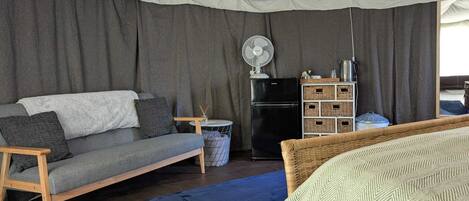 A cozy room with a grey sofa, a small round table, a black refrigerator, wicker baskets, and a bed with a patterned blanket.