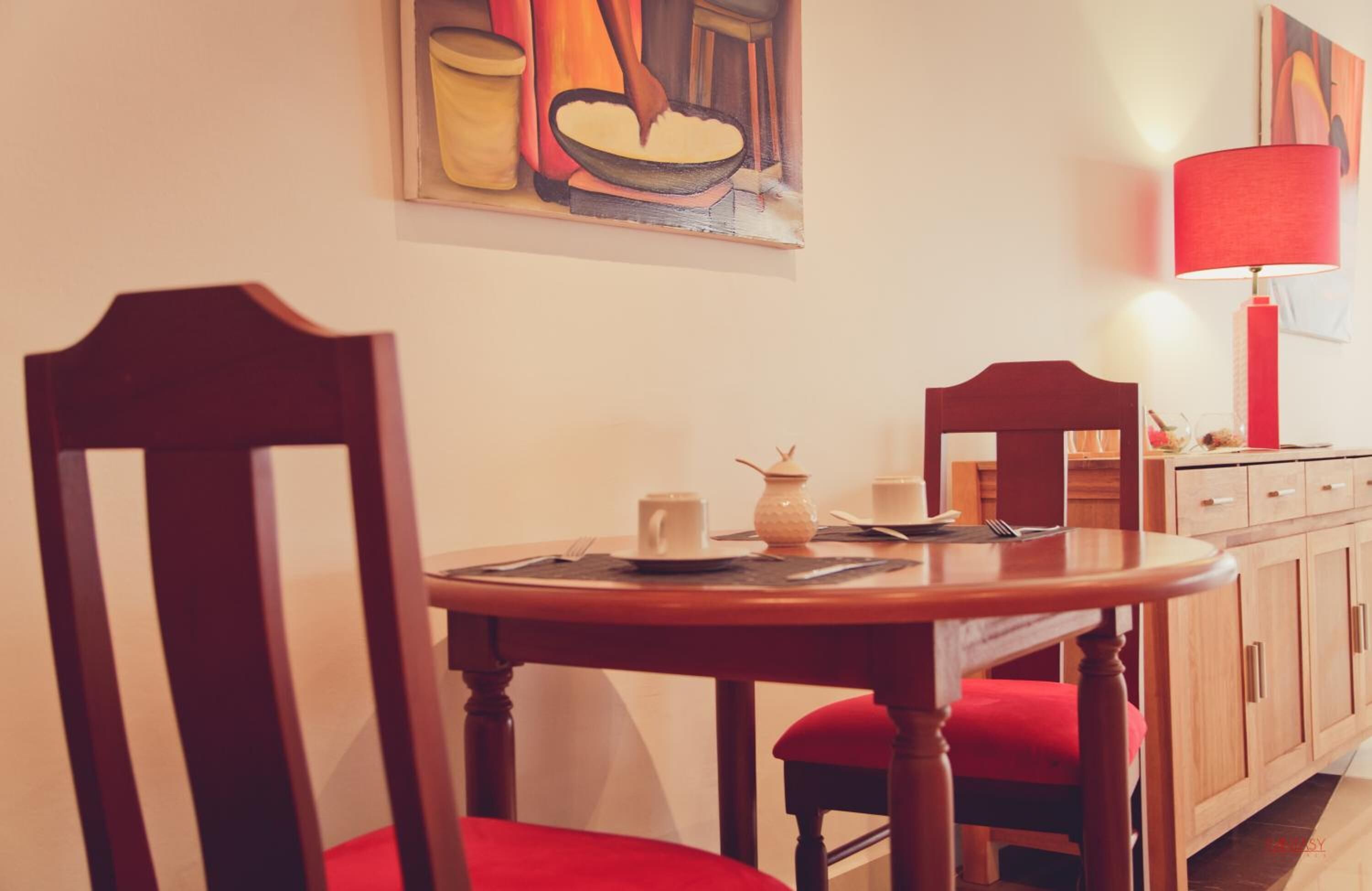 Un comedor con una mesa de madera, sillas rojas, una lámpara roja y un cuadro en la pared.