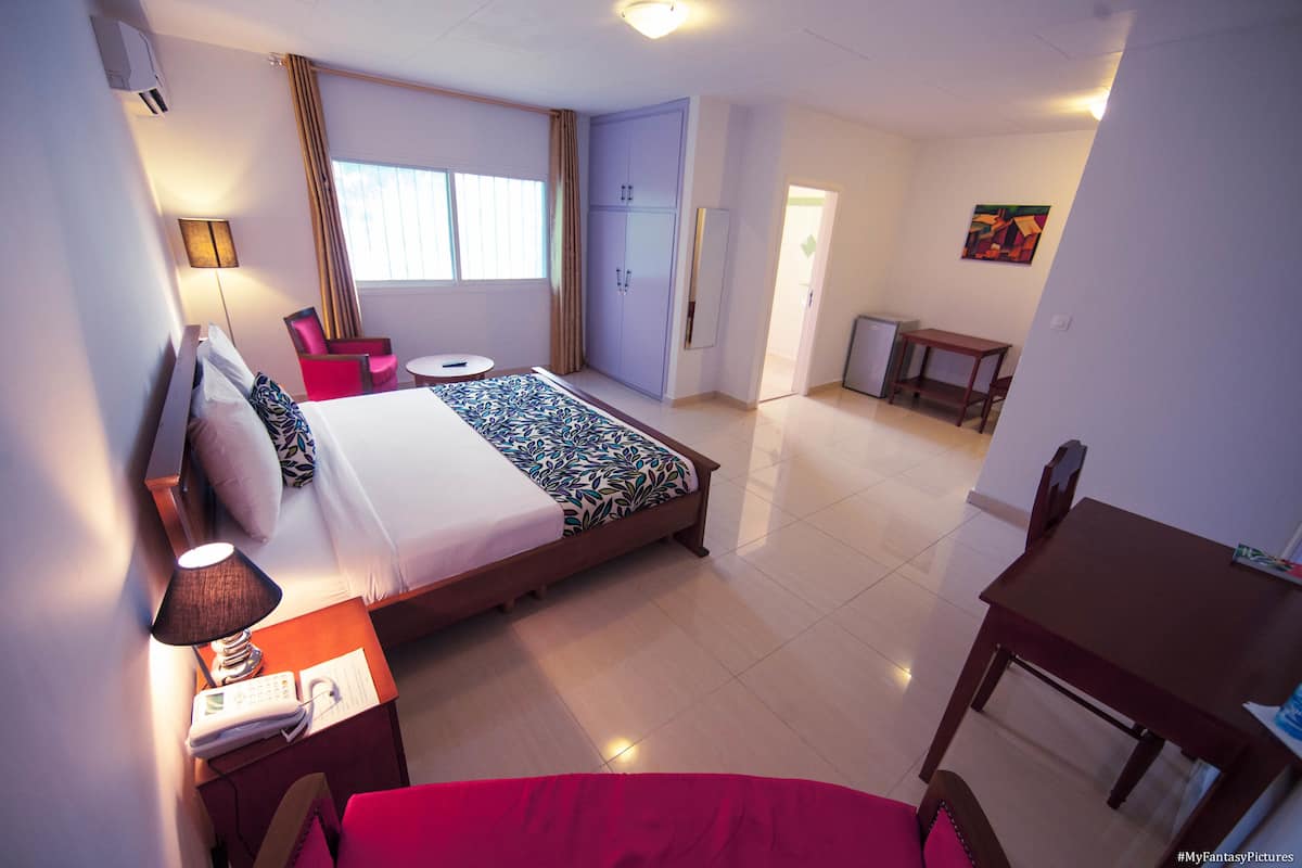A hotel room with a bed, a desk, a chair, a small table, a lamp, and a window with curtains.