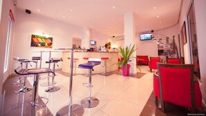 Un lobby moderno de hotel con mostrador de recepción, taburetes de bar, televisor y una planta grande.