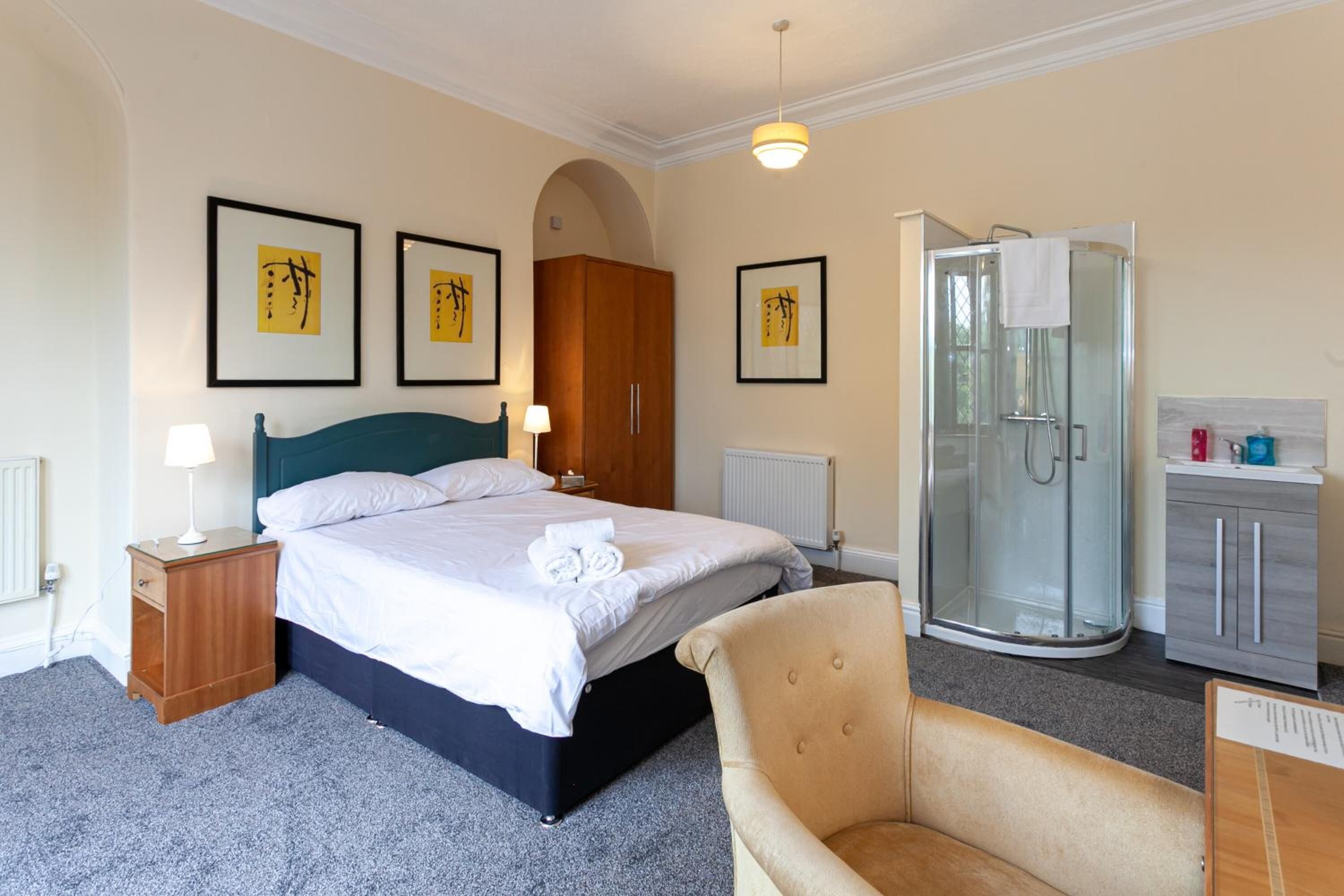 A hotel room with a bed, a chair, a shower, a wardrobe, and framed artwork on the wall.