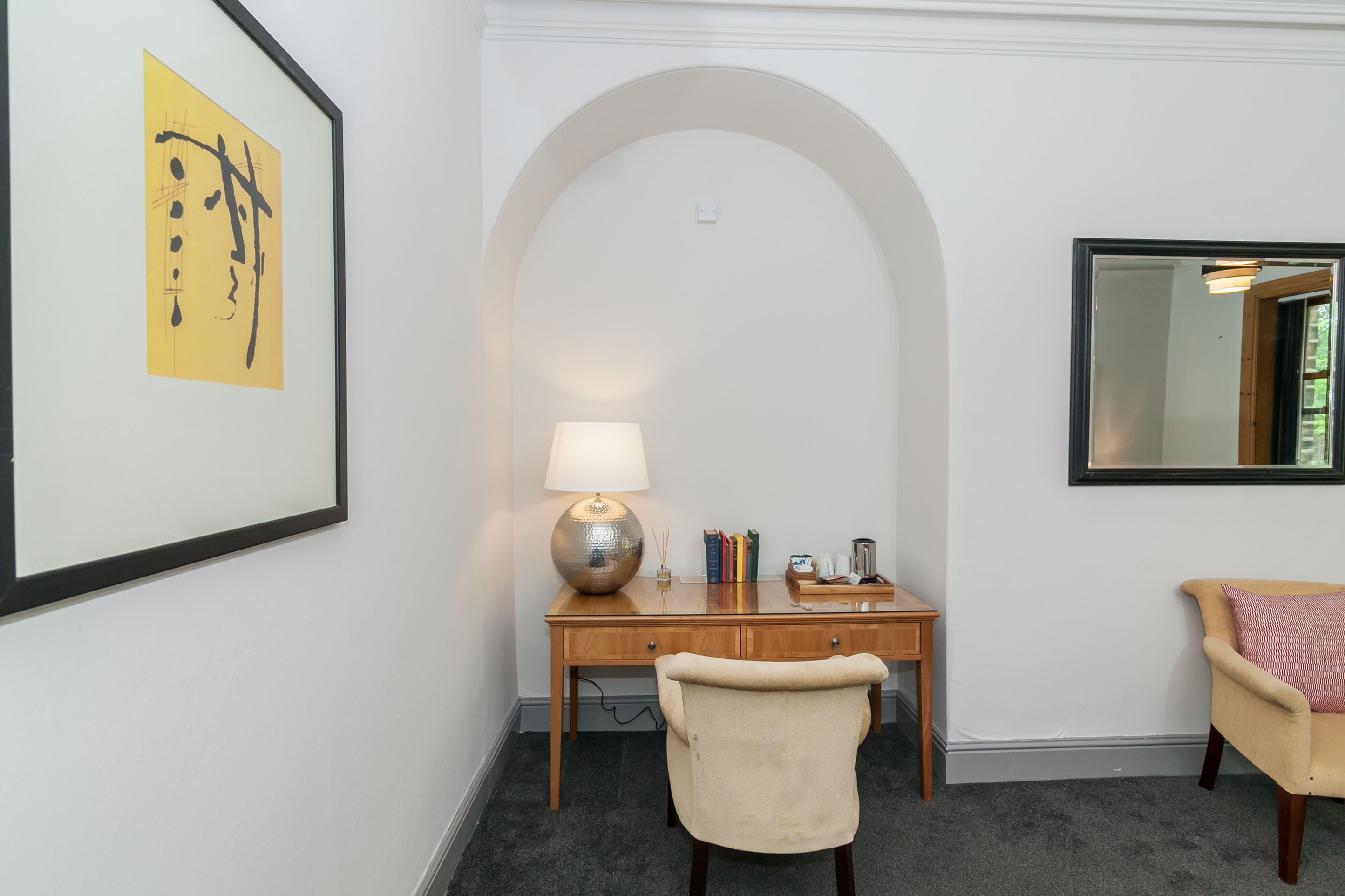 Une pièce comprenant un bureau, une chaise, une lampe, des livres et une œuvre d’art encadrée.