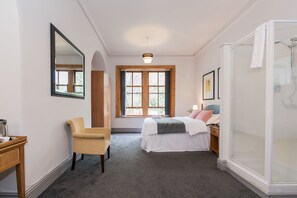 A bedroom with a bed, a chair, a desk, a window, and a shower.