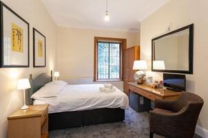 Habitación de hotel con cama, lámparas de noche, escritorio con ordenador, silla, espejo grande y obras de arte en las paredes.