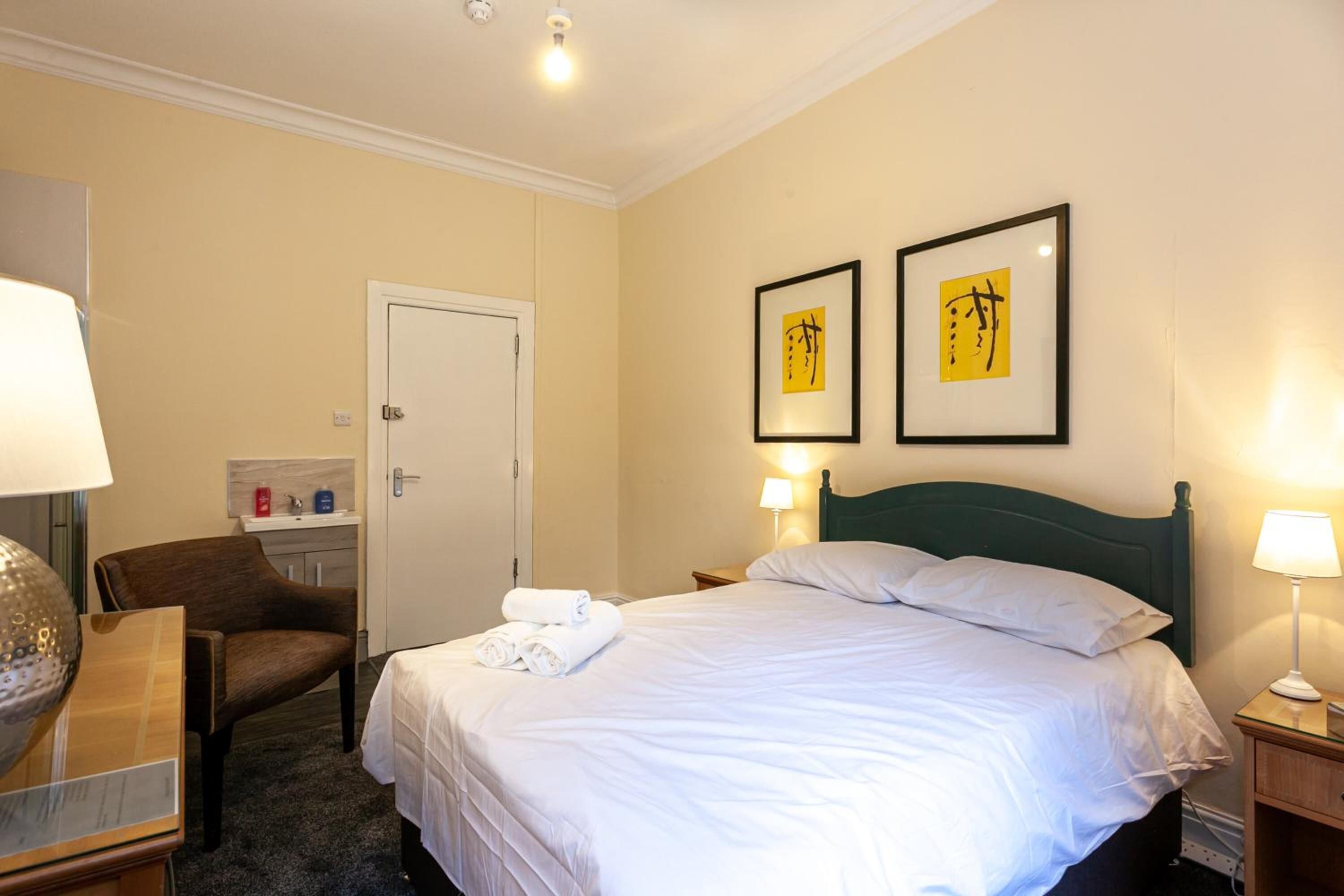 Ein Hotelzimmer mit einem Bett, einem Sessel, einem Schreibtisch, einer Lampe und zwei gerahmten Kunstwerken an der Wand.