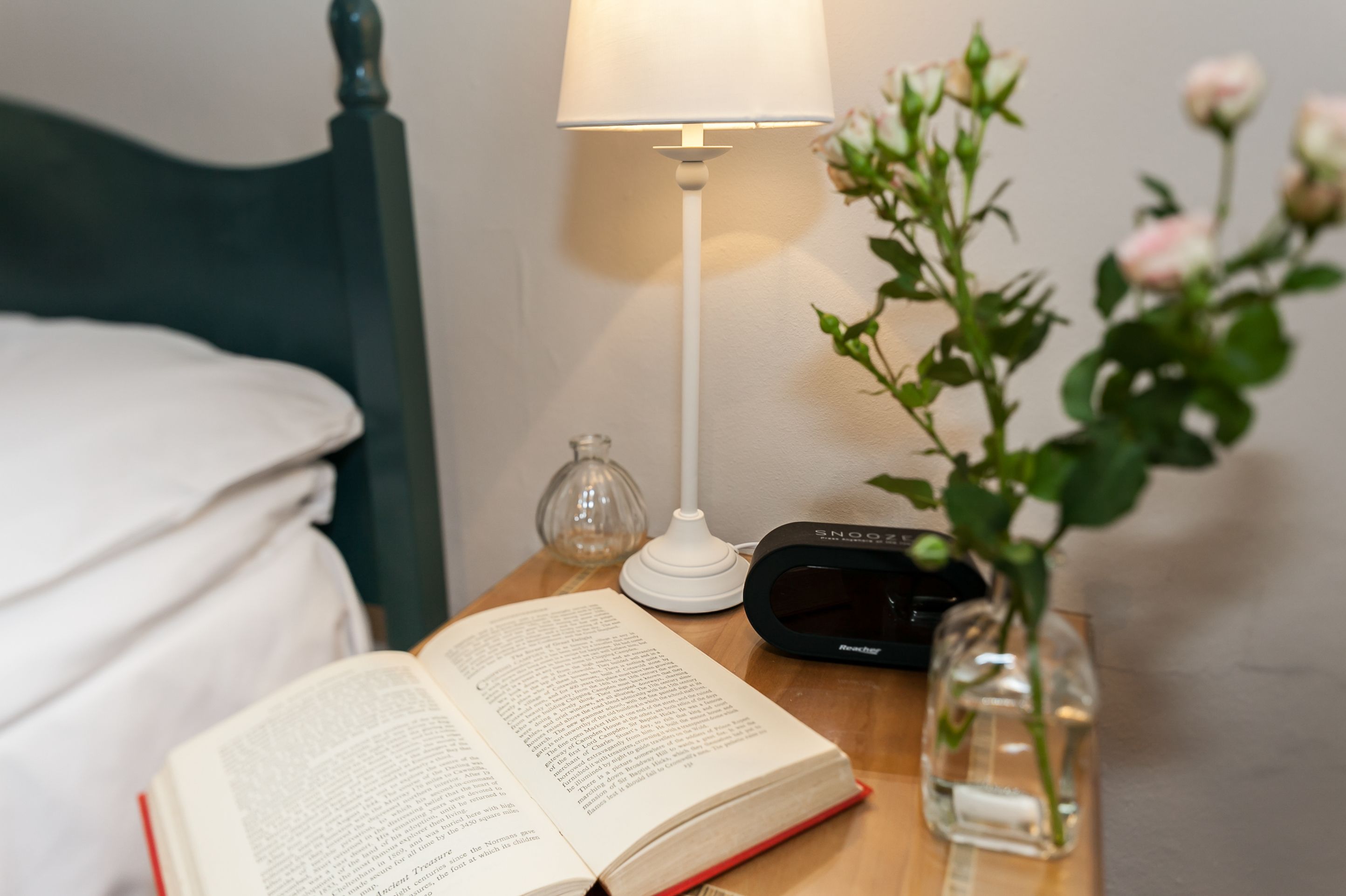 A bedside table with a book, a lamp, a clock, and a vase with a plant.