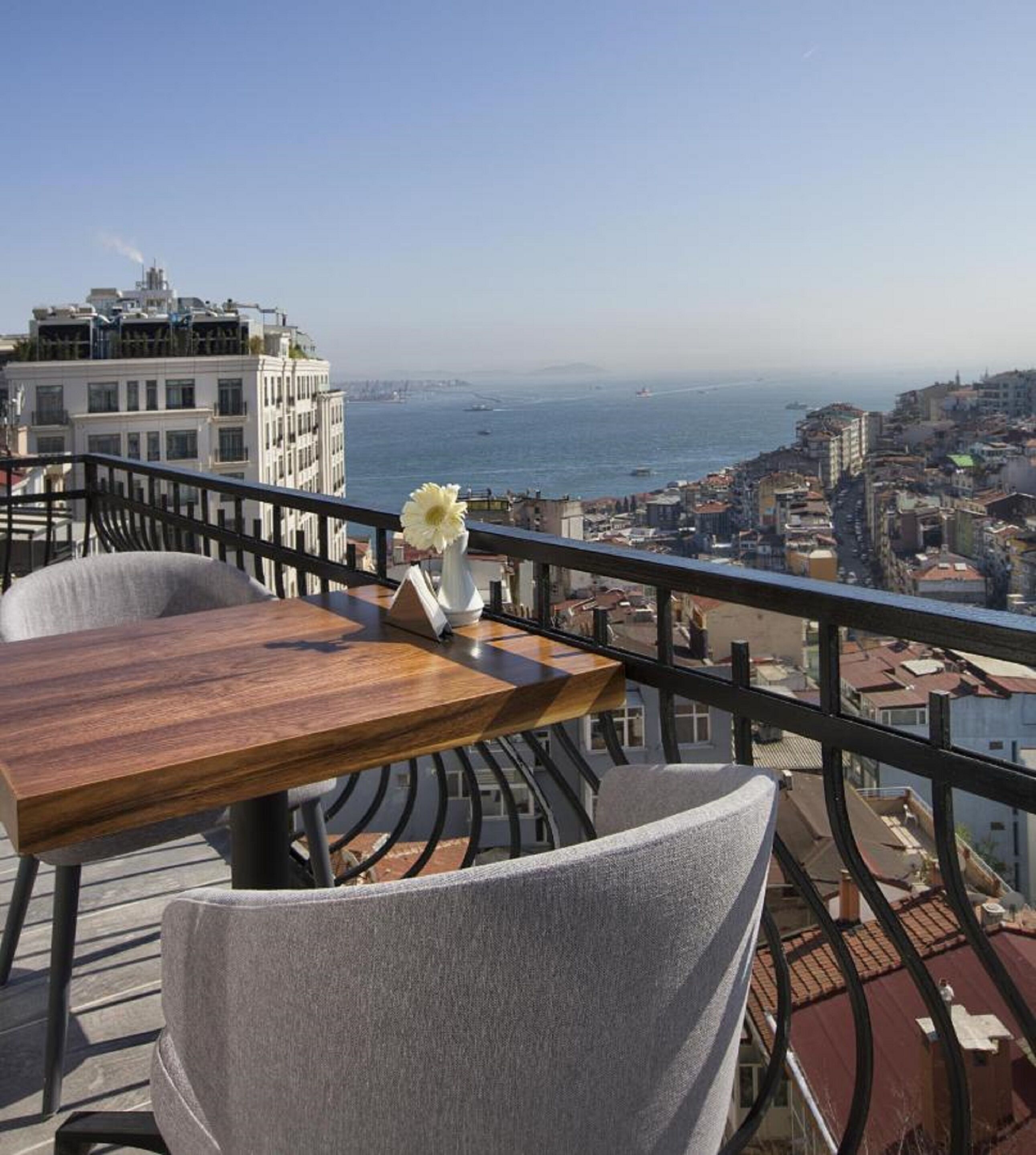Un balcon avec une table en bois et deux chaises, d’où l’on peut admirer un paysage urbain côtier avec ses bâtiments et la mer.
