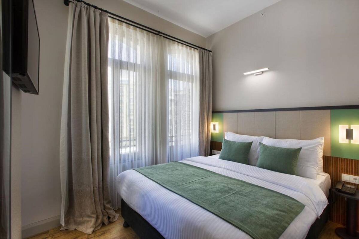 A hotel room with a large bed, a green throw blanket, a nightstand with a phone, and a window with sheer curtains.