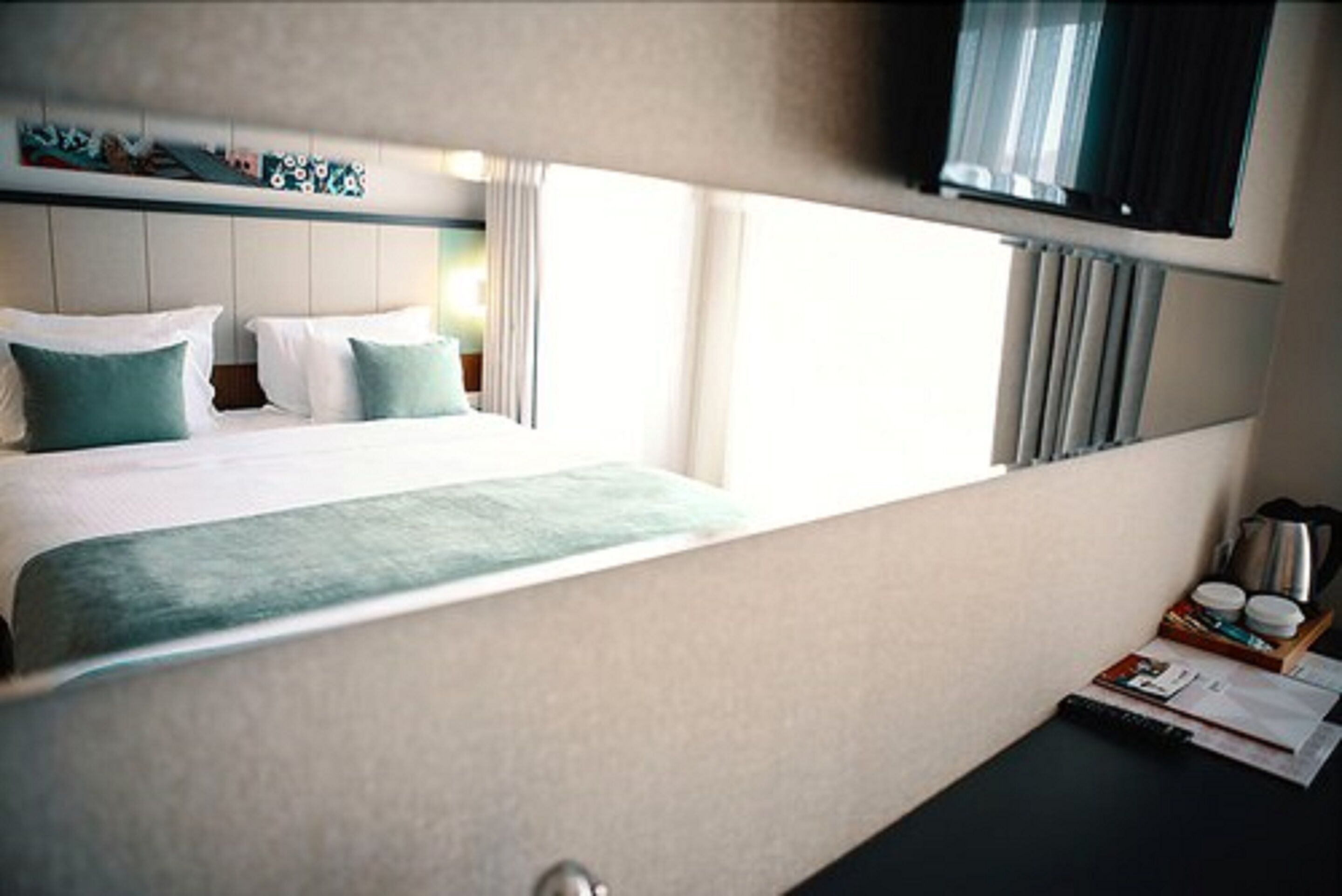 A hotel room with a bed, a flat-screen TV, a small table with a kettle, and a window with curtains.