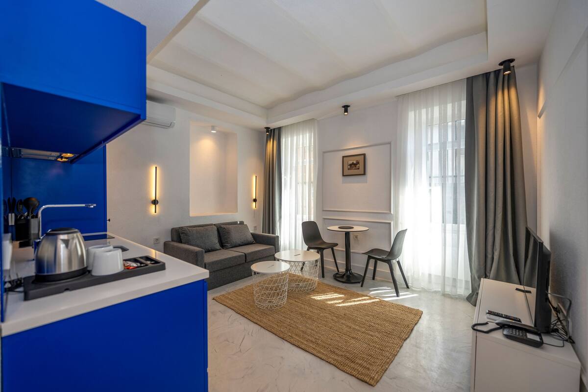A modern hotel room with a kitchenette, a grey sofa, a small dining table, and a TV.