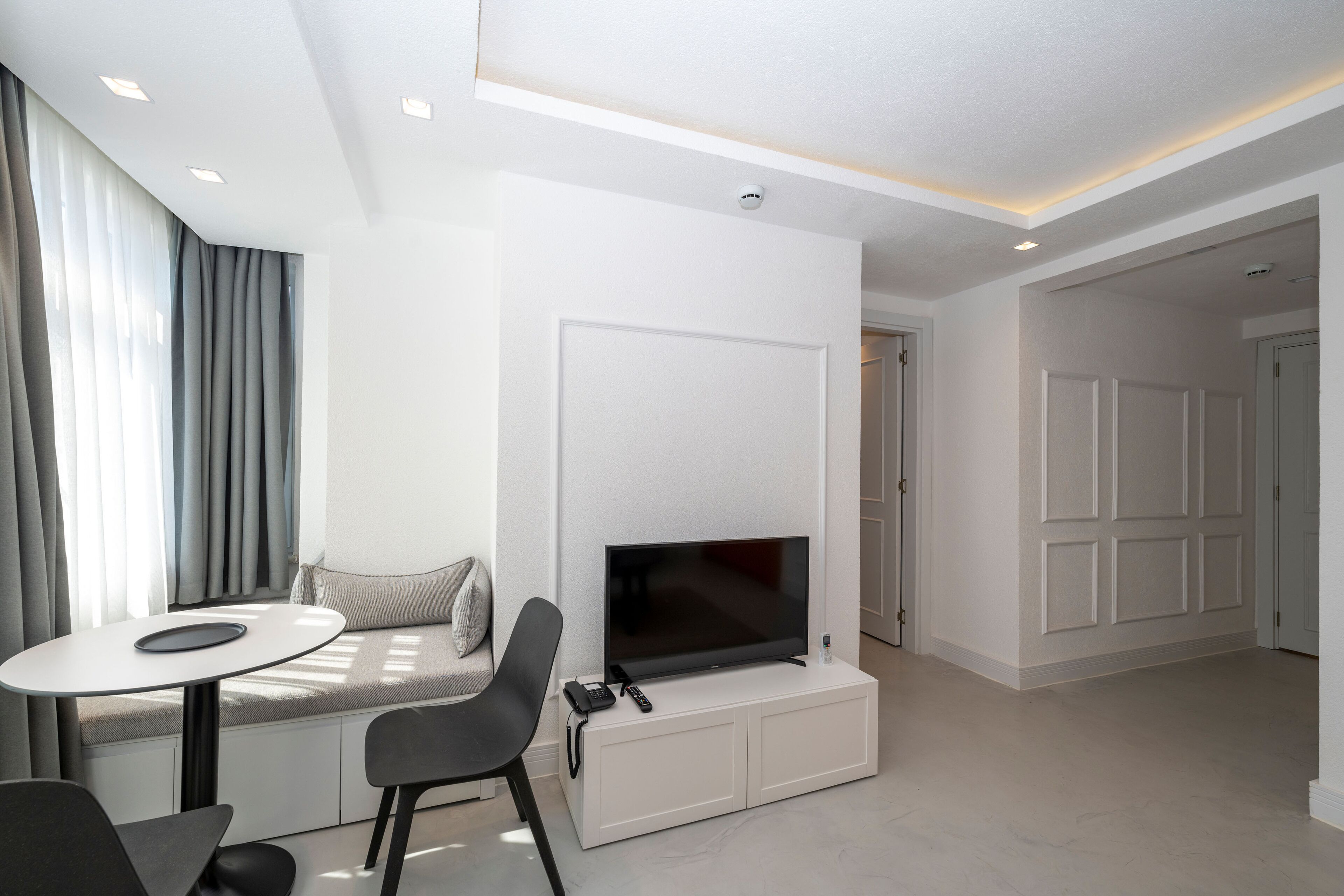 Uma sala de estar moderna com uma televisão de tela plana fixada em um armário branco, uma mesa de jantar com cadeiras e um sofá com um almofada.