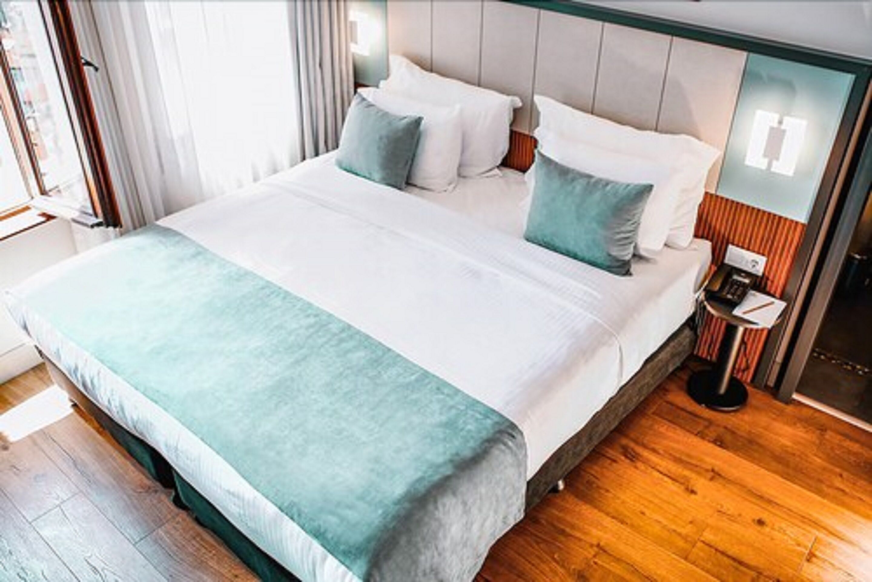 A modern hotel room with a large bed, a bedside table, and a view of the city through the window.