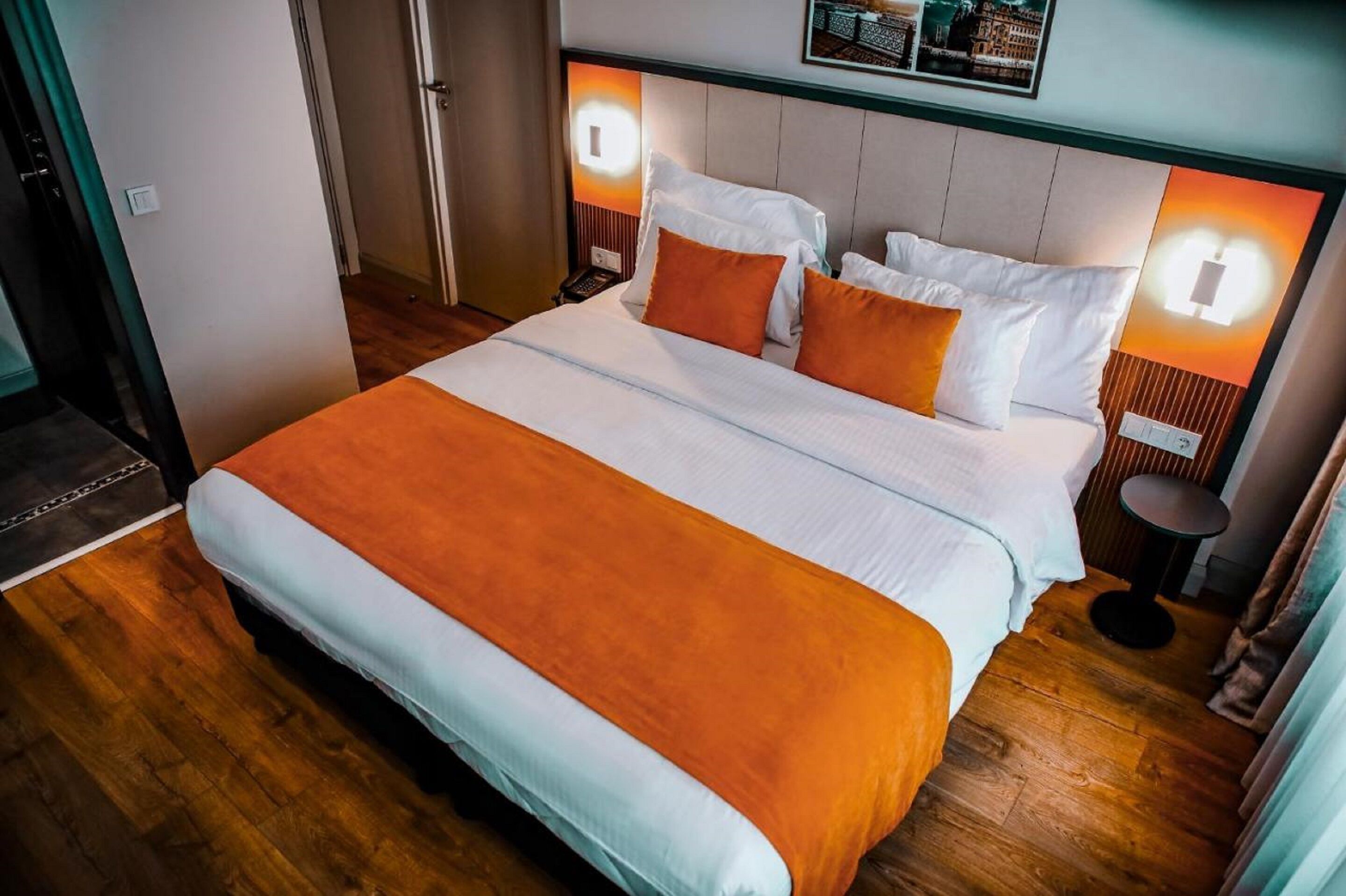 Una cama bien hecha con ropa de cama blanca y naranja, suelo de madera y una mesita redonda al lado.