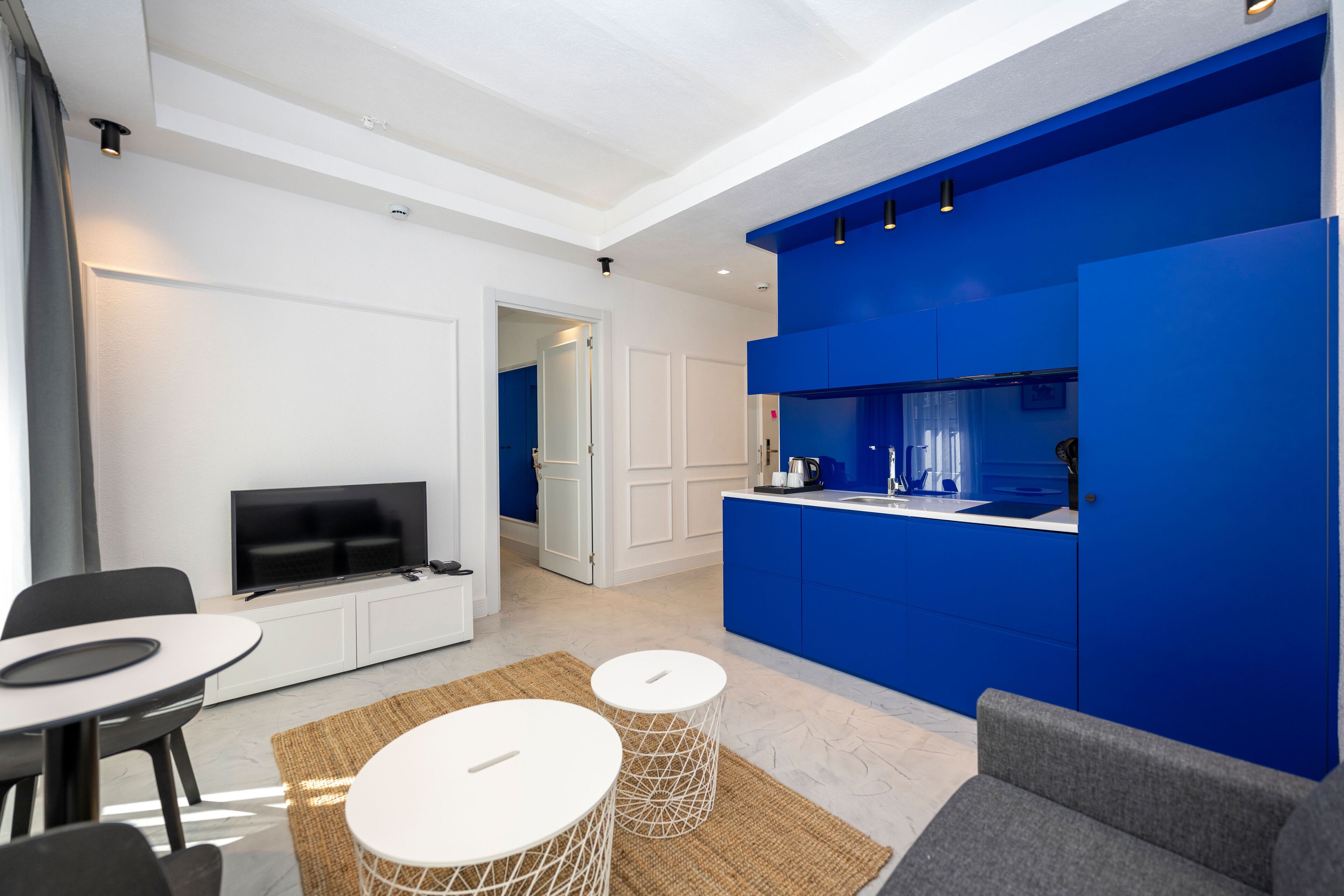 A modern living room with a flat-screen TV, a dining area with a round table, and a kitchen with blue cabinets.