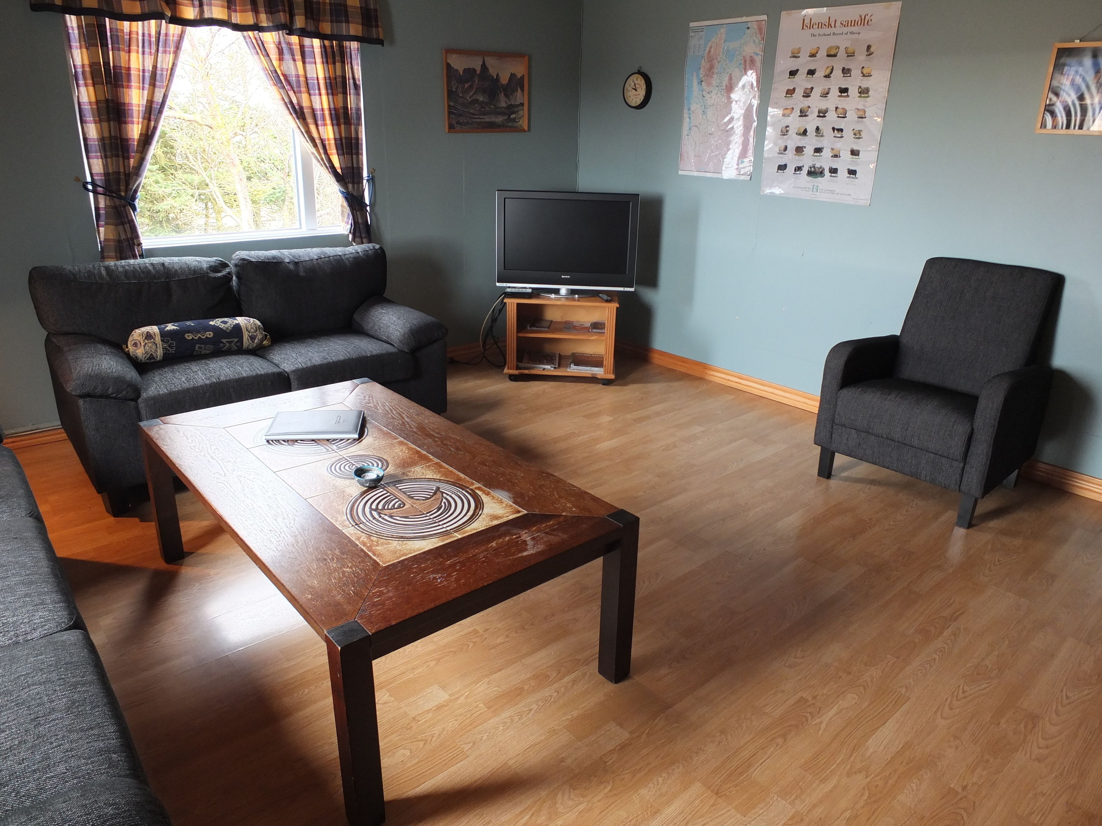 A living room with a sofa, armchair, coffee table, TV, and a wall with a clock and posters.