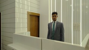 A man in a grey suit standing in a modern interior with white paneling and a wooden door. - Hotel Muskan (Mathura)