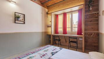 Una habitación con una cama, un escritorio con una silla, una ventana con cortinas y una planta colgante.