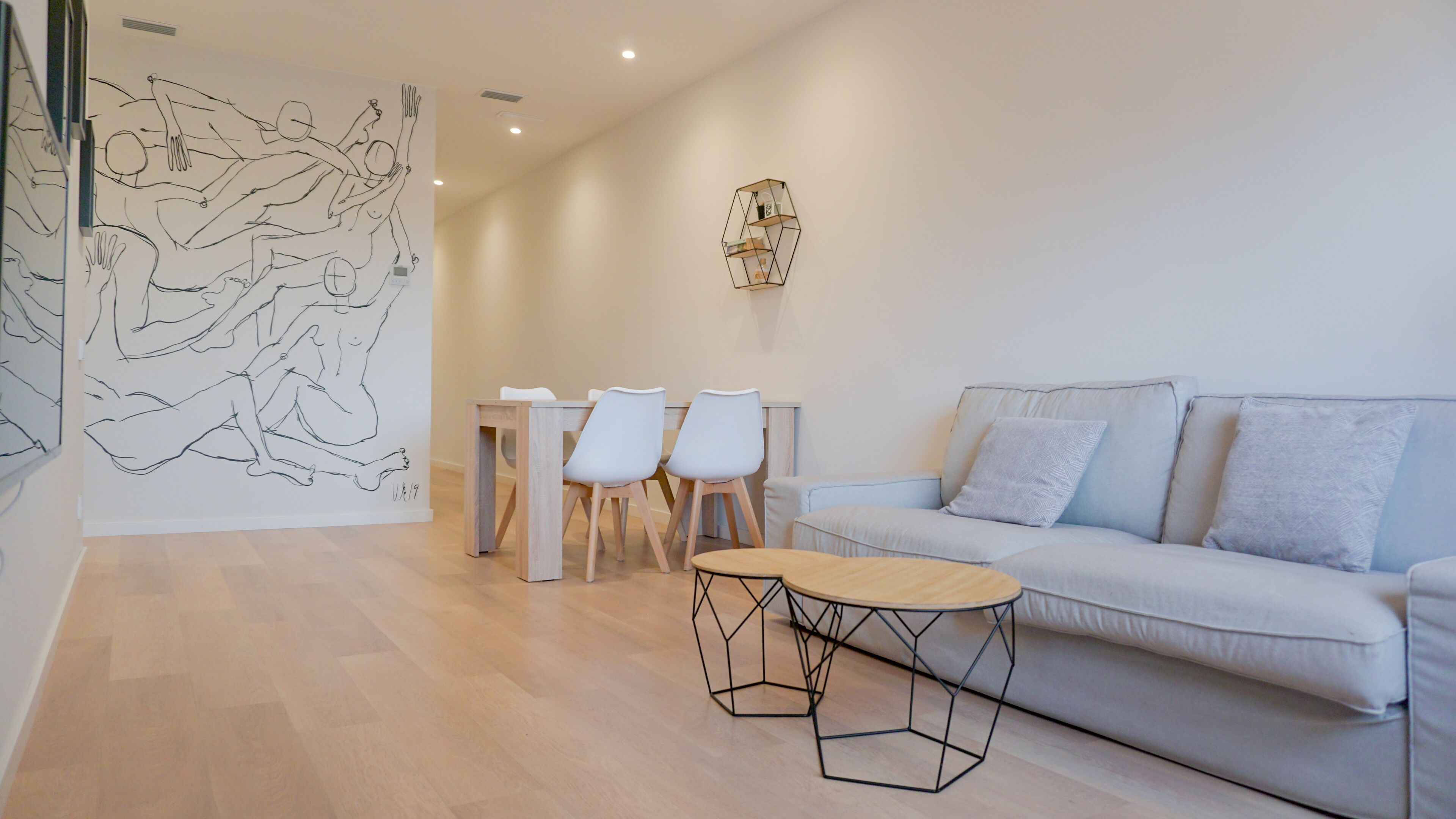Una sala de estar moderna con un sofá de color gris claro, dos sillas blancas, una mesa de centro de madera y una obra de arte geométrica en la pared.