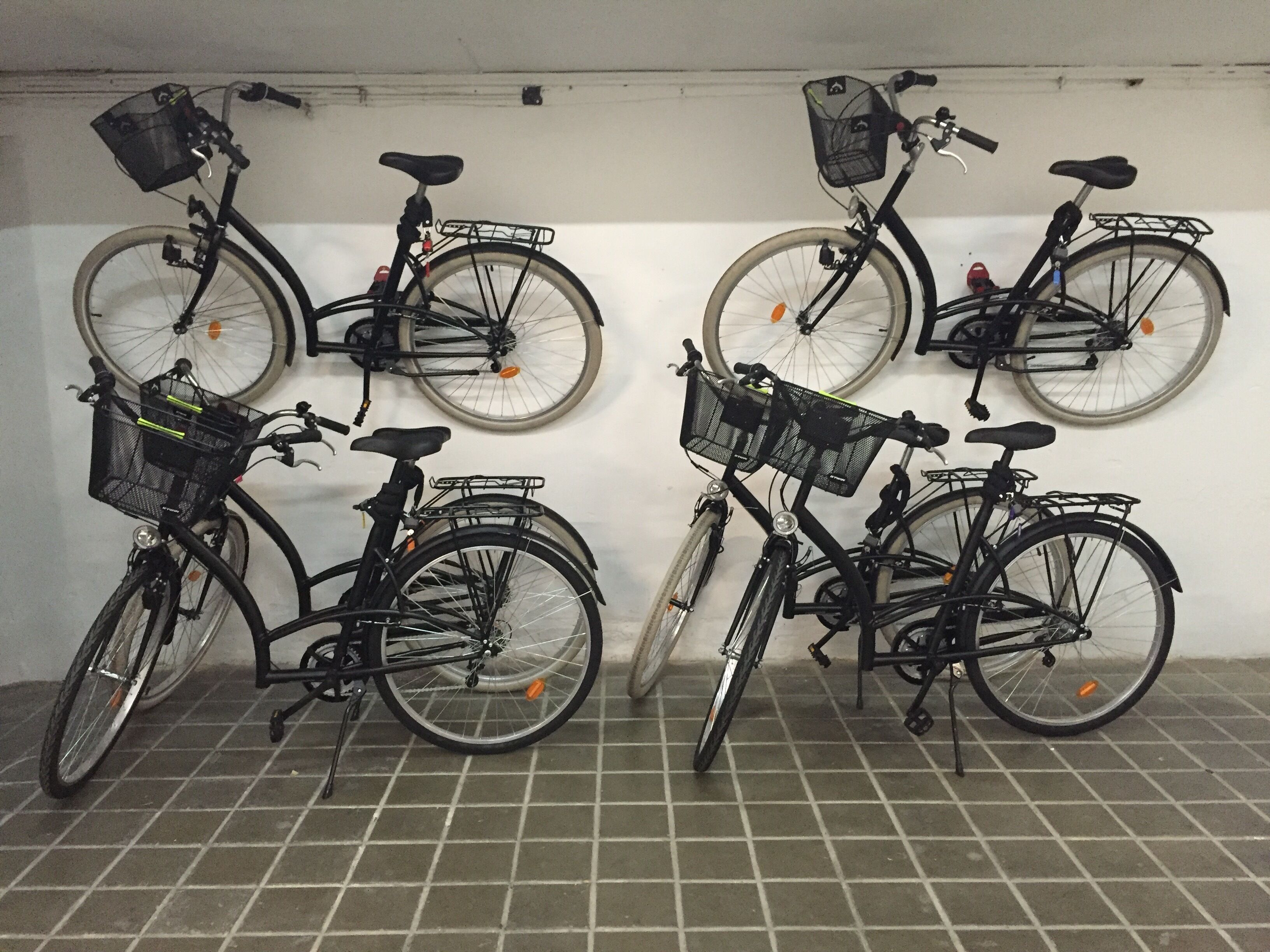 A row of bicycles with baskets and kickstands, parked against a wall in a tiled room.