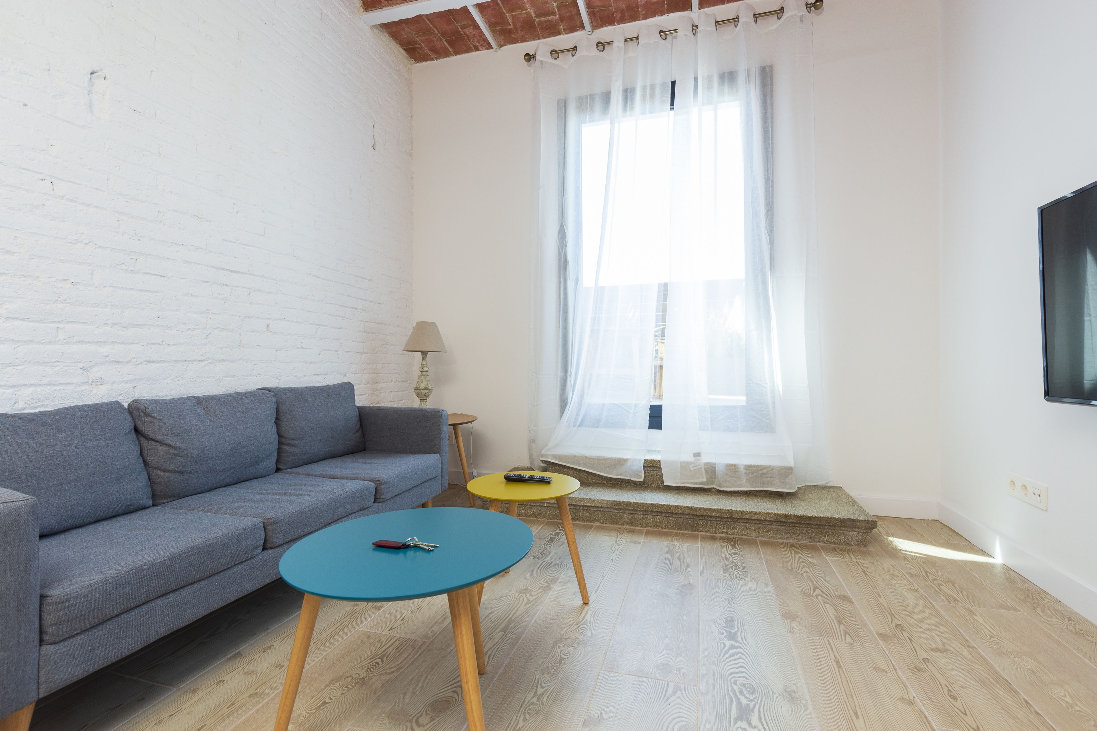 Un salon avec un canapé gris, une table bleue, une petite table jaune et une fenêtre ornée de rideaux légers.