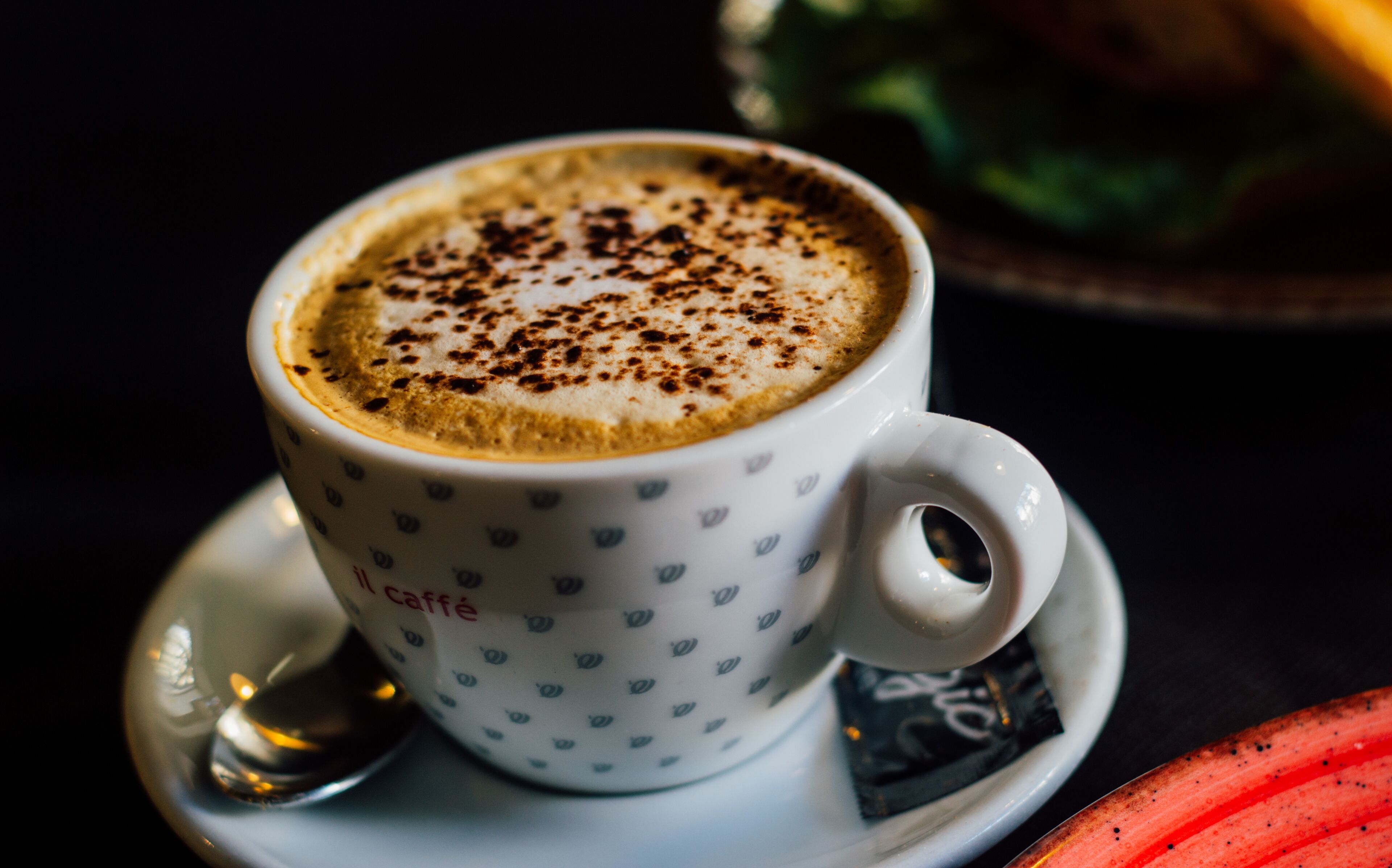 Una taza de café con platito y cuchara, y una vista parcial de un plato con comida.