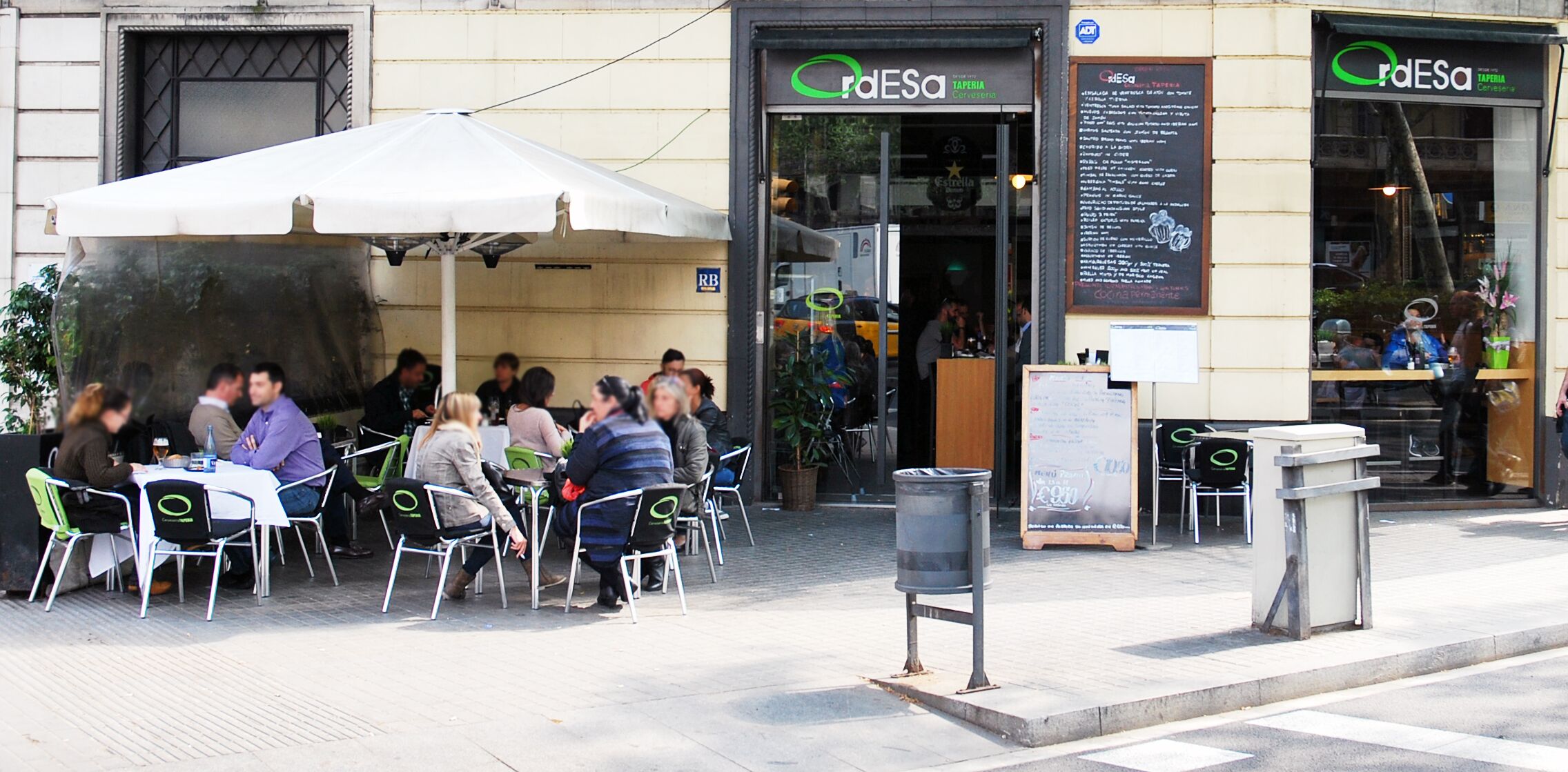 An outdoor café with tables and chairs, a large umbrella, and a menu board outside a restaurant named 'dESA'.