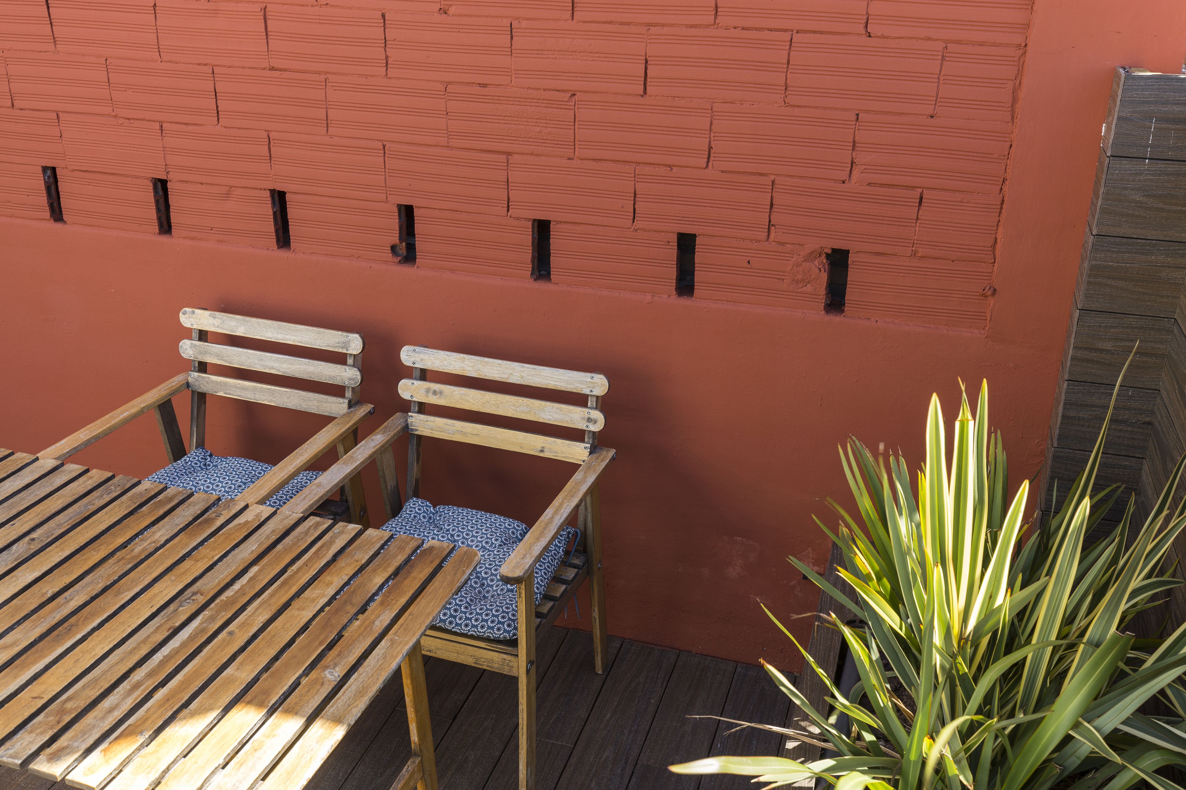 Een houten tafel met twee stoelen en een gekussend kussen, geplaatst tegen een bakstenen muur met een raam.