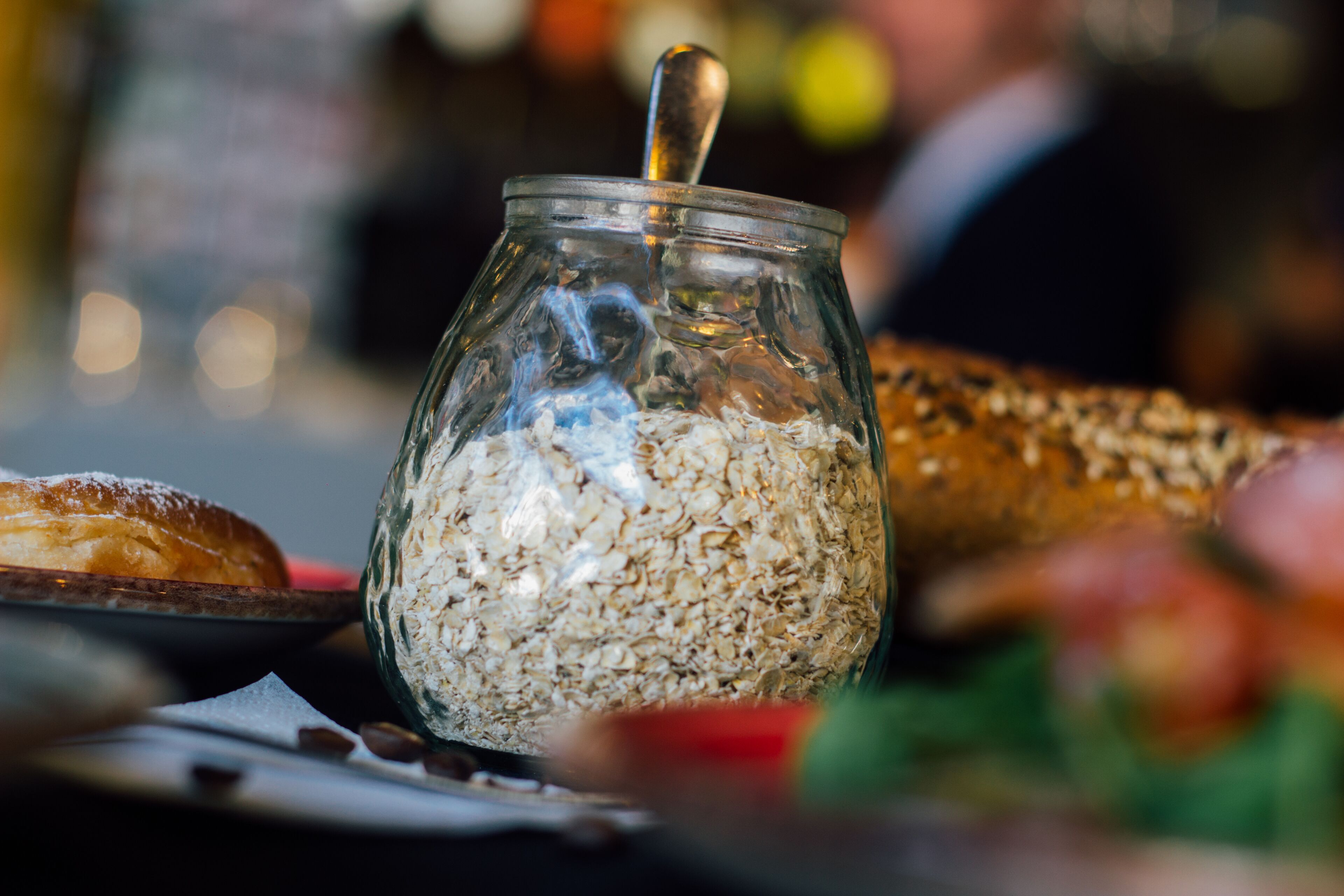 Un tarro de vidrio lleno de avena y una cuchara en su interior, colocado sobre una mesa con diferentes alimentos a su alrededor.