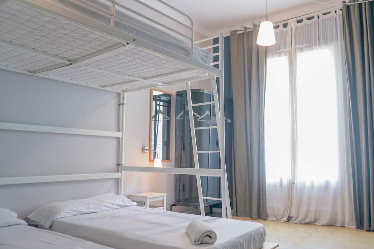 Quarto de hotel com beliches, uma mesinha, uma escada, uma luminária pendente e uma janela grande com cortinas.