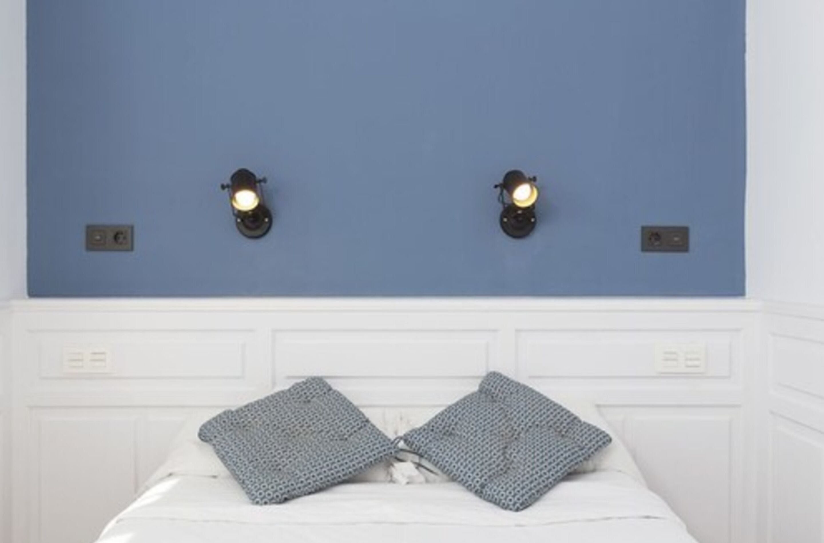 A bedroom with a blue wall, two wall-mounted light fixtures, and a bed with two patterned pillows.