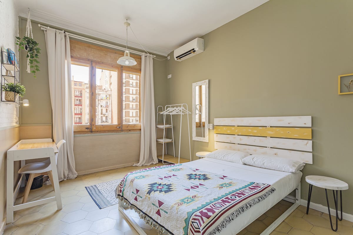 Quarto com cama, escrivaninha, cadeira, planta pendente e janela com cortinas.