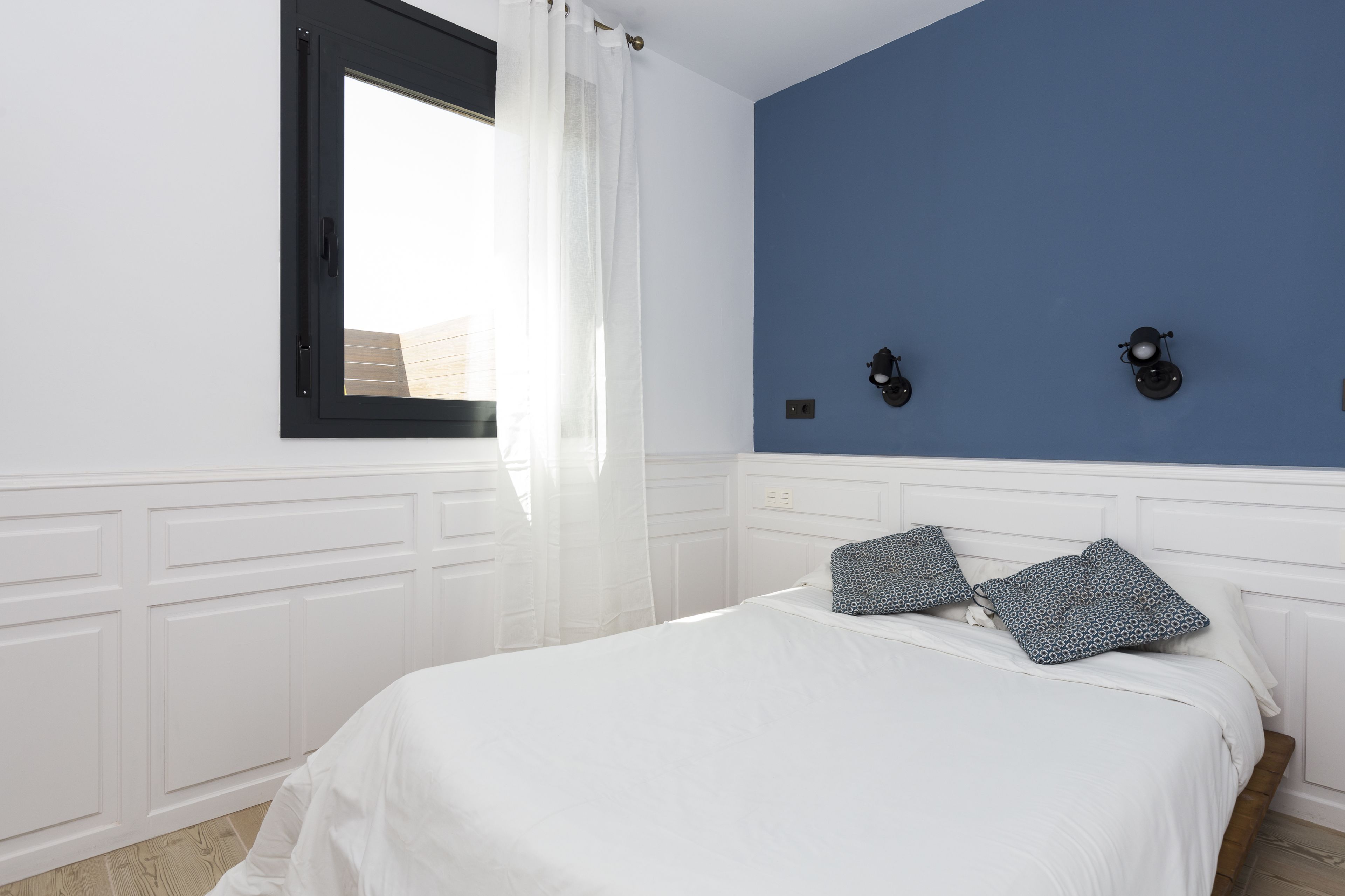 Ein Schlafzimmer mit einem Bett, weißen Vorhängen, einem Fenster und einer blauen Wand mit Wandleuchten.