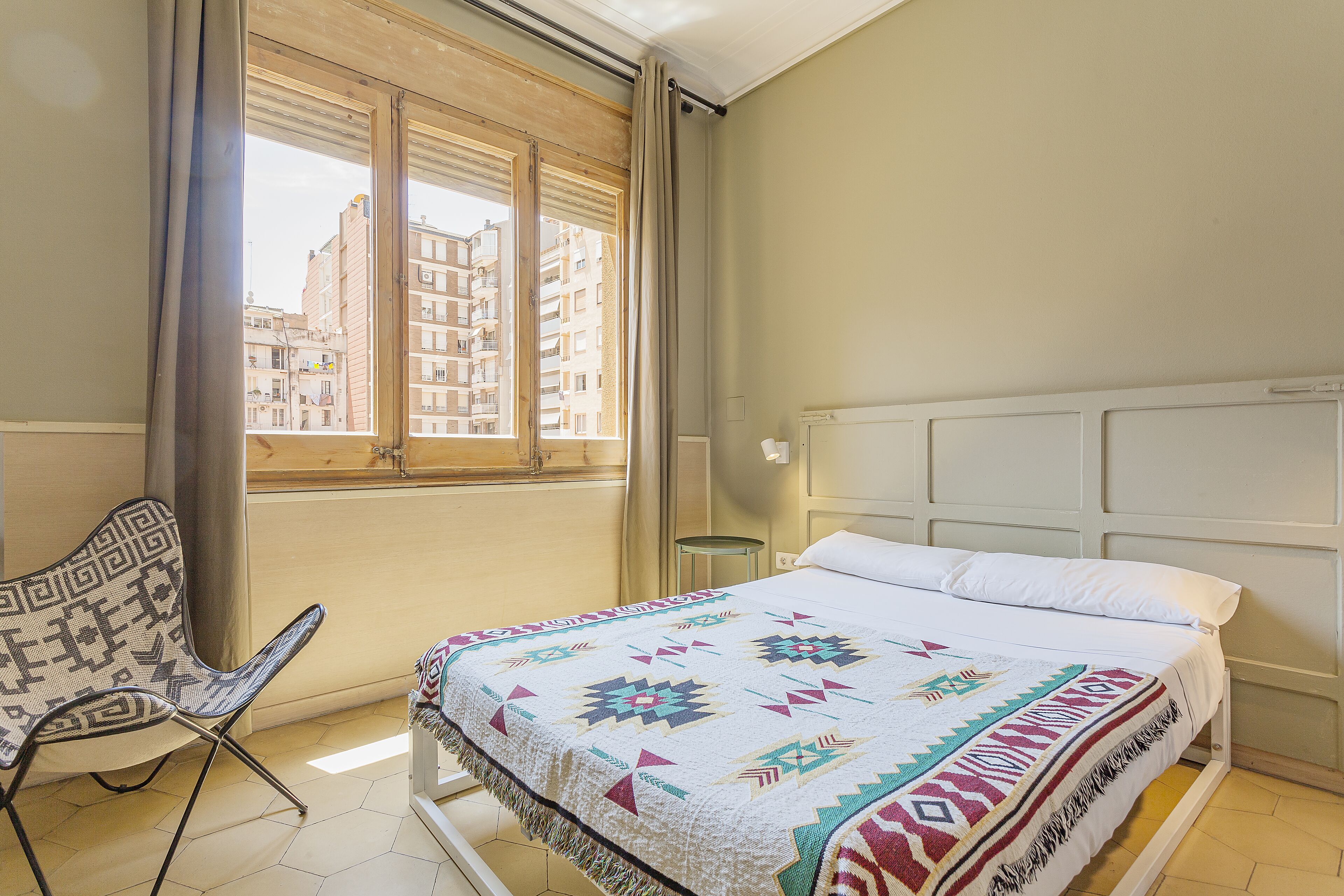 A bedroom with a bed, a chair, a window with curtains, and a view of buildings outside.