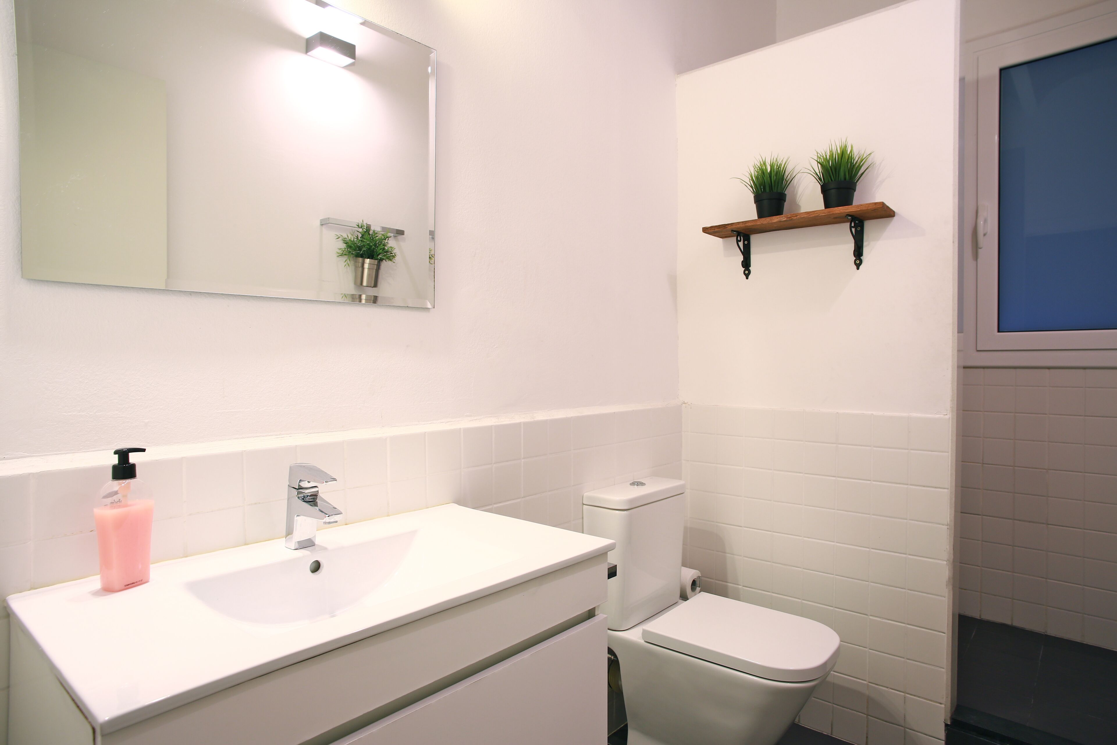 Een badkamer met een wit wastafelmeubel, toilet, spiegel en twee planten in potten op een plank.