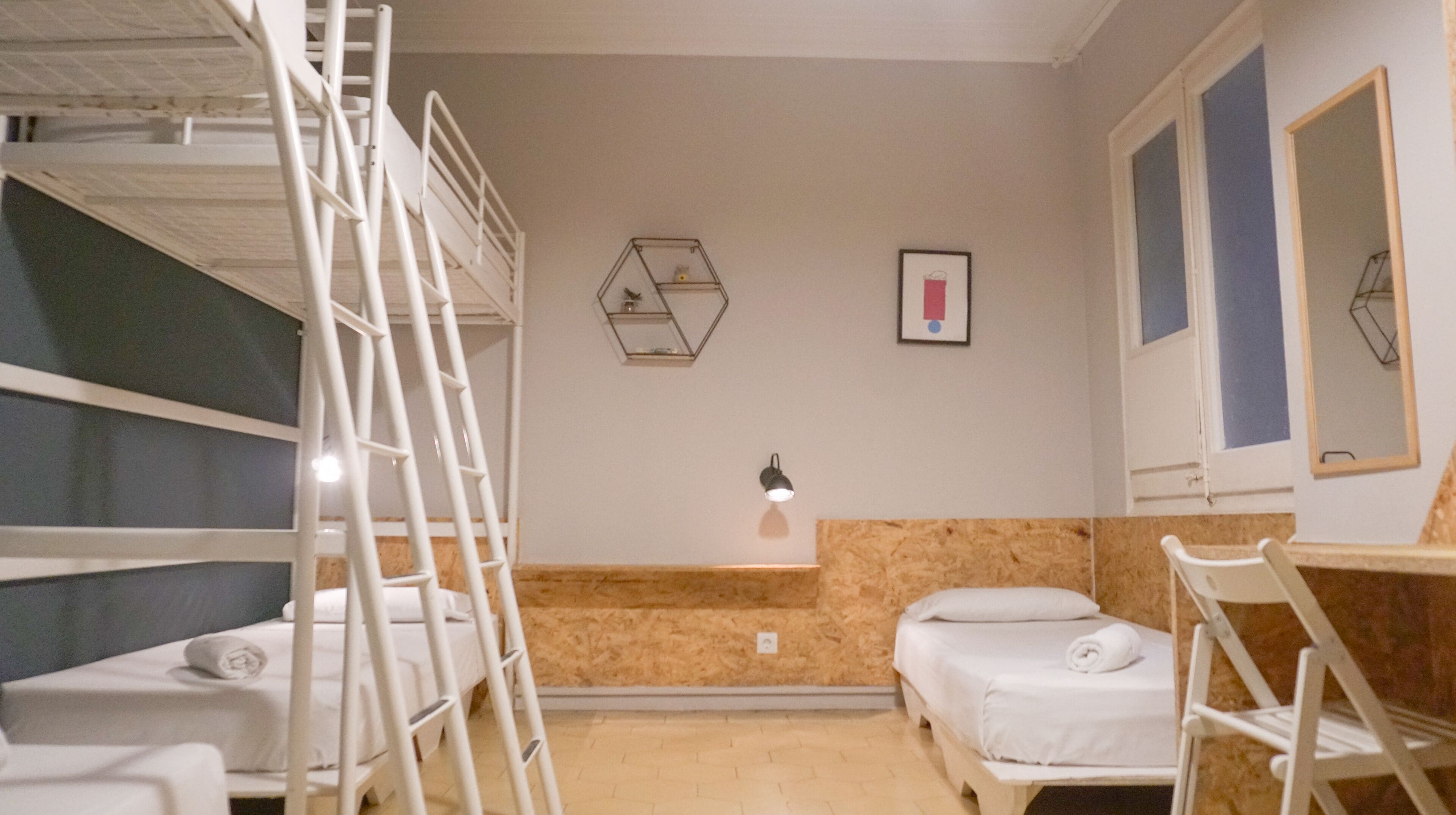 A dormitory room with bunk beds, a desk, a chair, a small bedside table, and a wall-mounted lamp.
