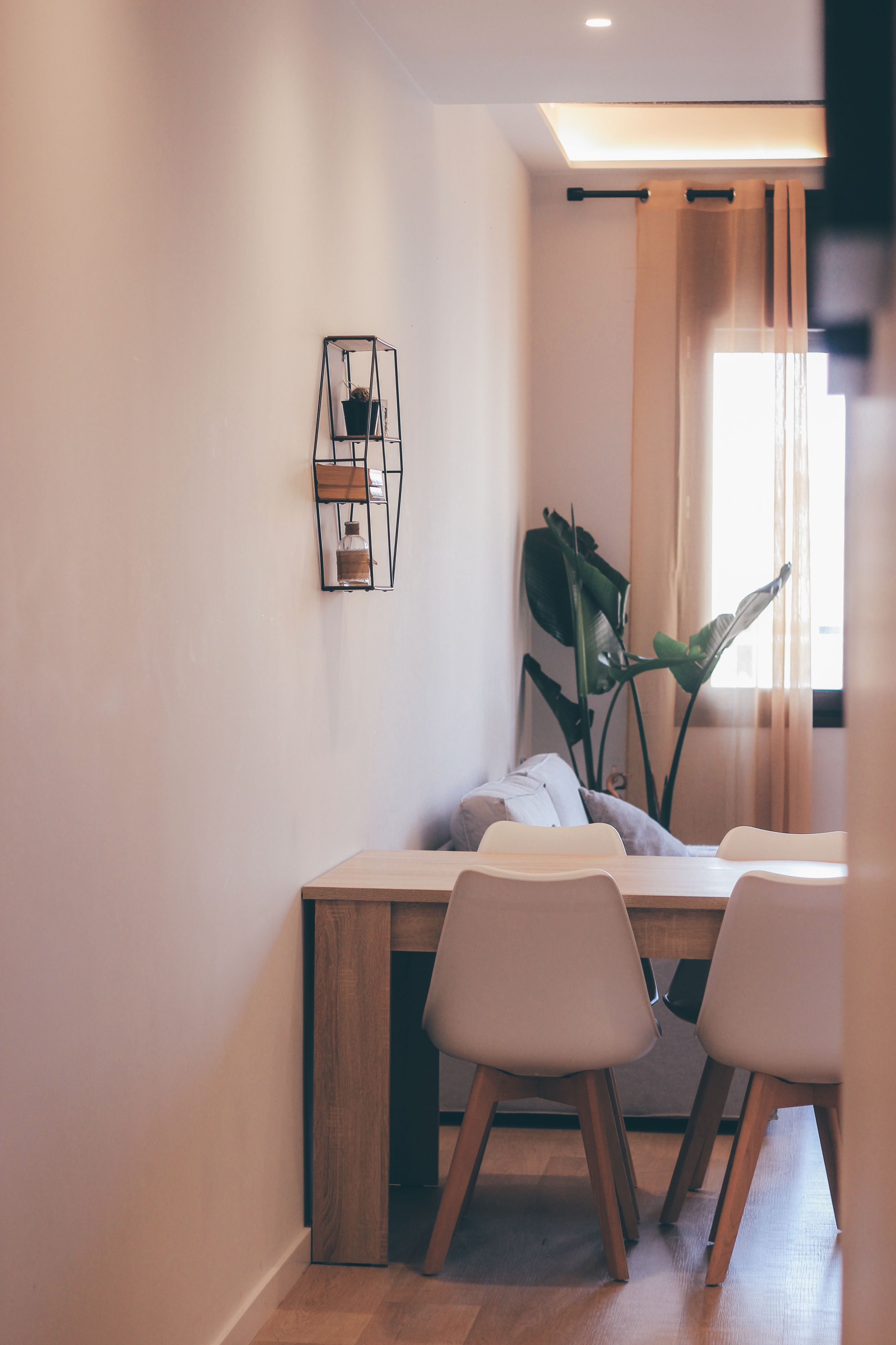 A modern dining area with a wooden table, two chairs, a potted plant, and a wall-mounted light fixture.