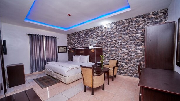 A hotel room with a bed, a desk, two chairs, a television, and a stone-patterned wall. - EEMJM Hotels and Suites Limited (Uyo)