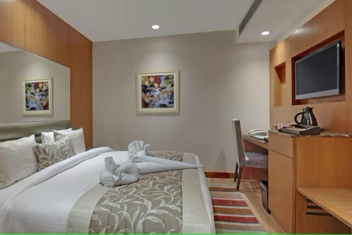 Quarto de hotel com cama, escrivaninha, cadeira, televisão e obras de arte nas paredes.
