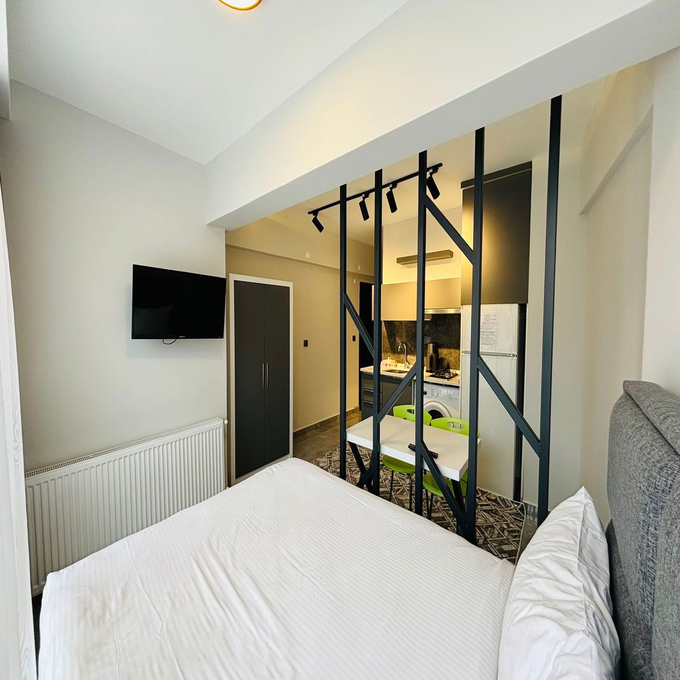 A hotel room with a bed, a TV mounted on the wall, a kitchenette, and a bathroom visible through a partition.