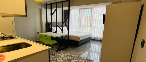 Une chambre d’hôtel compacte comprenant un coin cuisine, un lit avec un cadre noir, un petit bureau et un tapis à motifs.