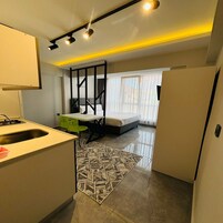 Habitación de hotel compacta con kitchenette, cama con estructura negra, escritorio pequeño y alfombra estampada.