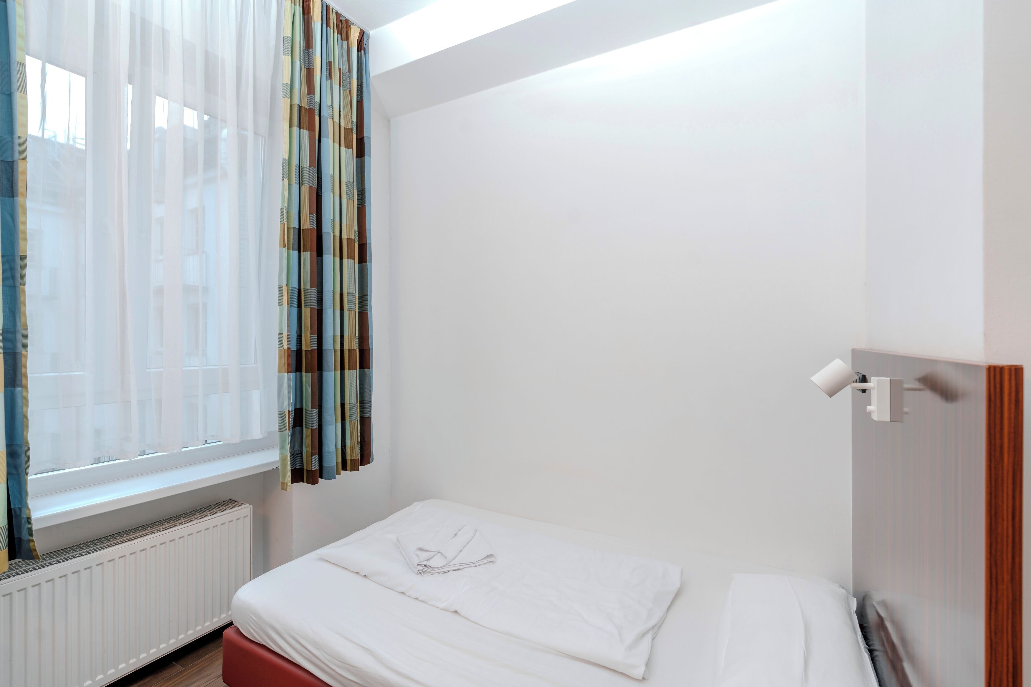 Una cama pequeña y bien hecha en una habitación de paredes blancas y una ventana con cortinas traslúcidas.