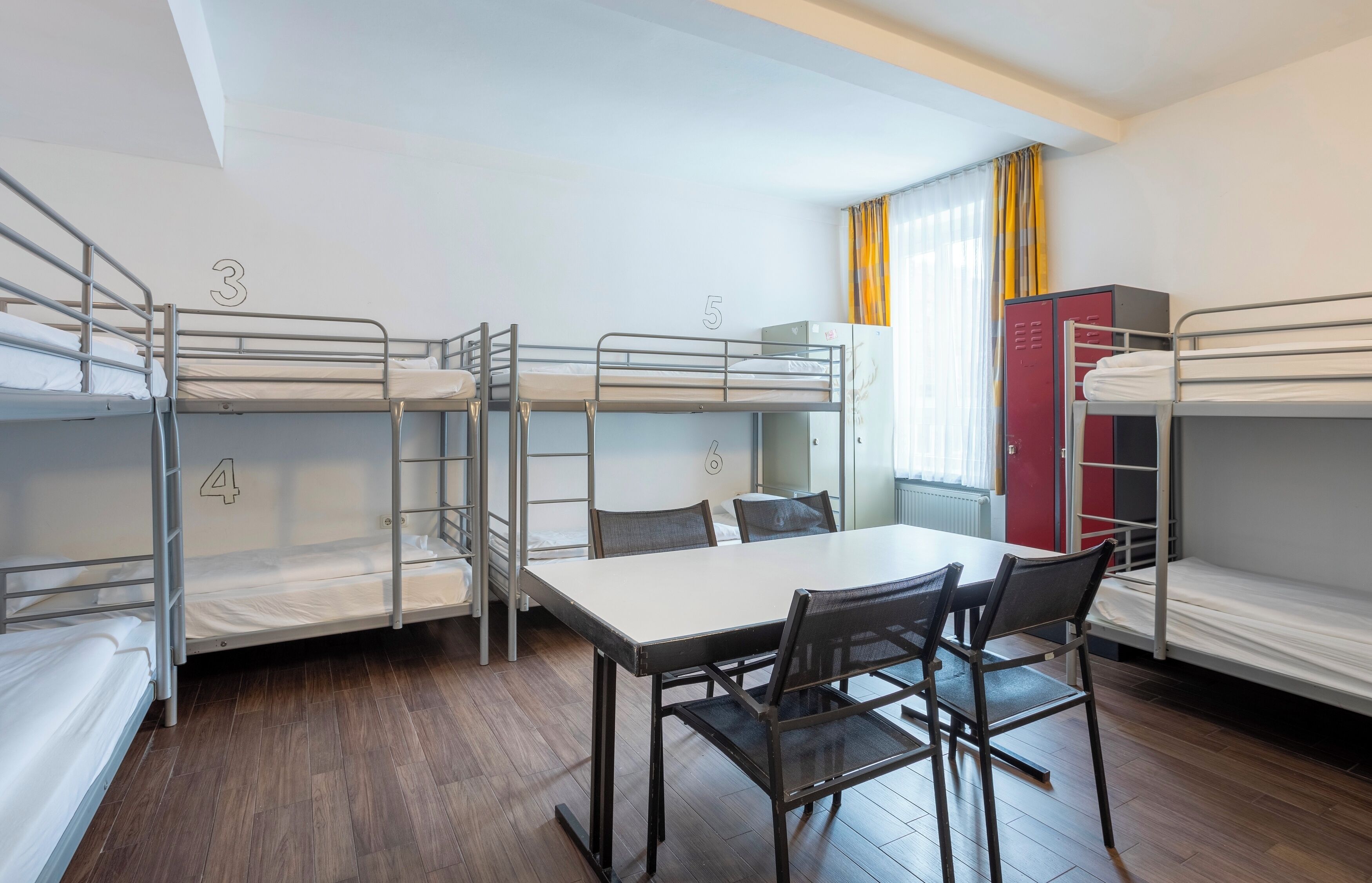 Habitación tipo dormitorio con literas numeradas 3, 4 y 5, una mesa con sillas y un armario rojo.