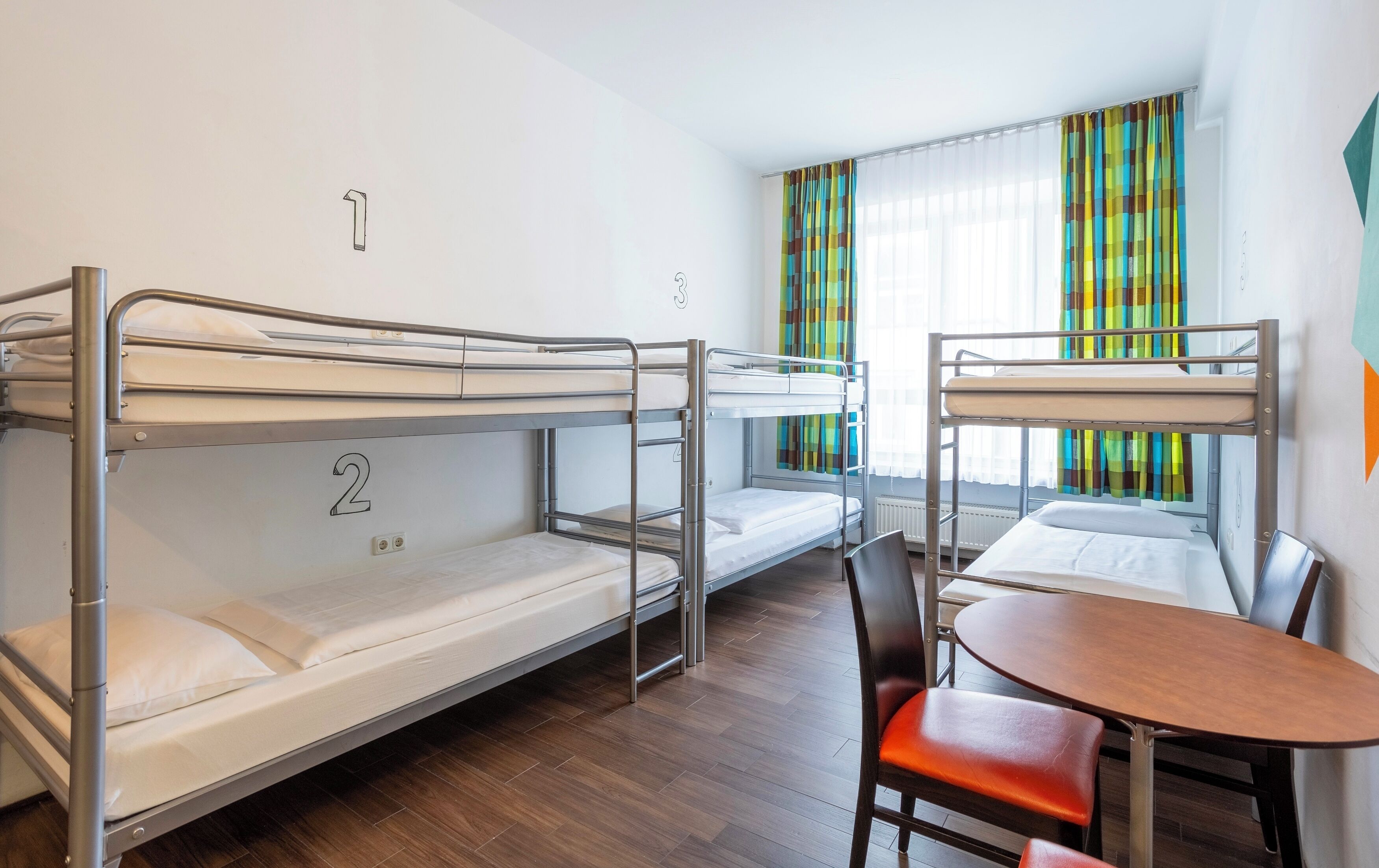 A dormitory room with bunk beds, a wooden floor, a small table, and colorful curtains.
